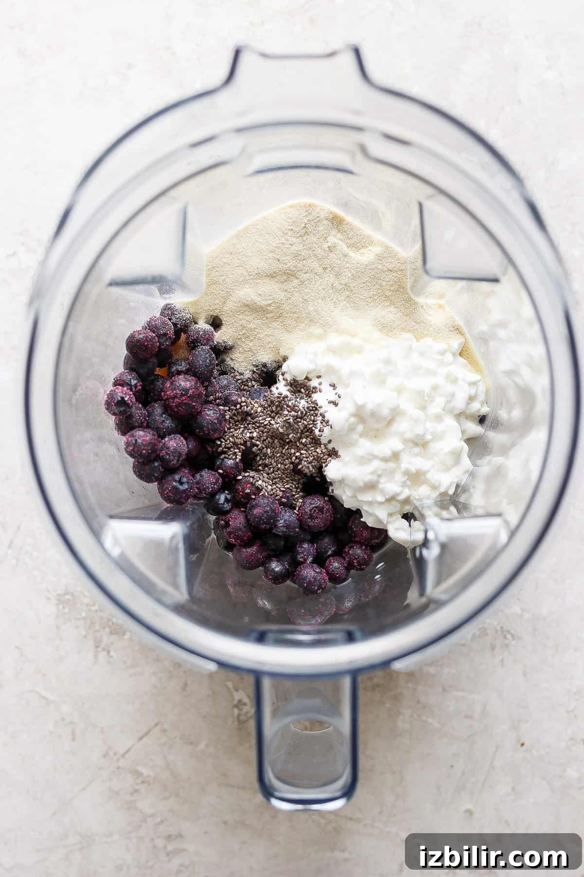 Top view of a blender containing blueberries, yogurt, chia seeds, and protein powder on a light surface.