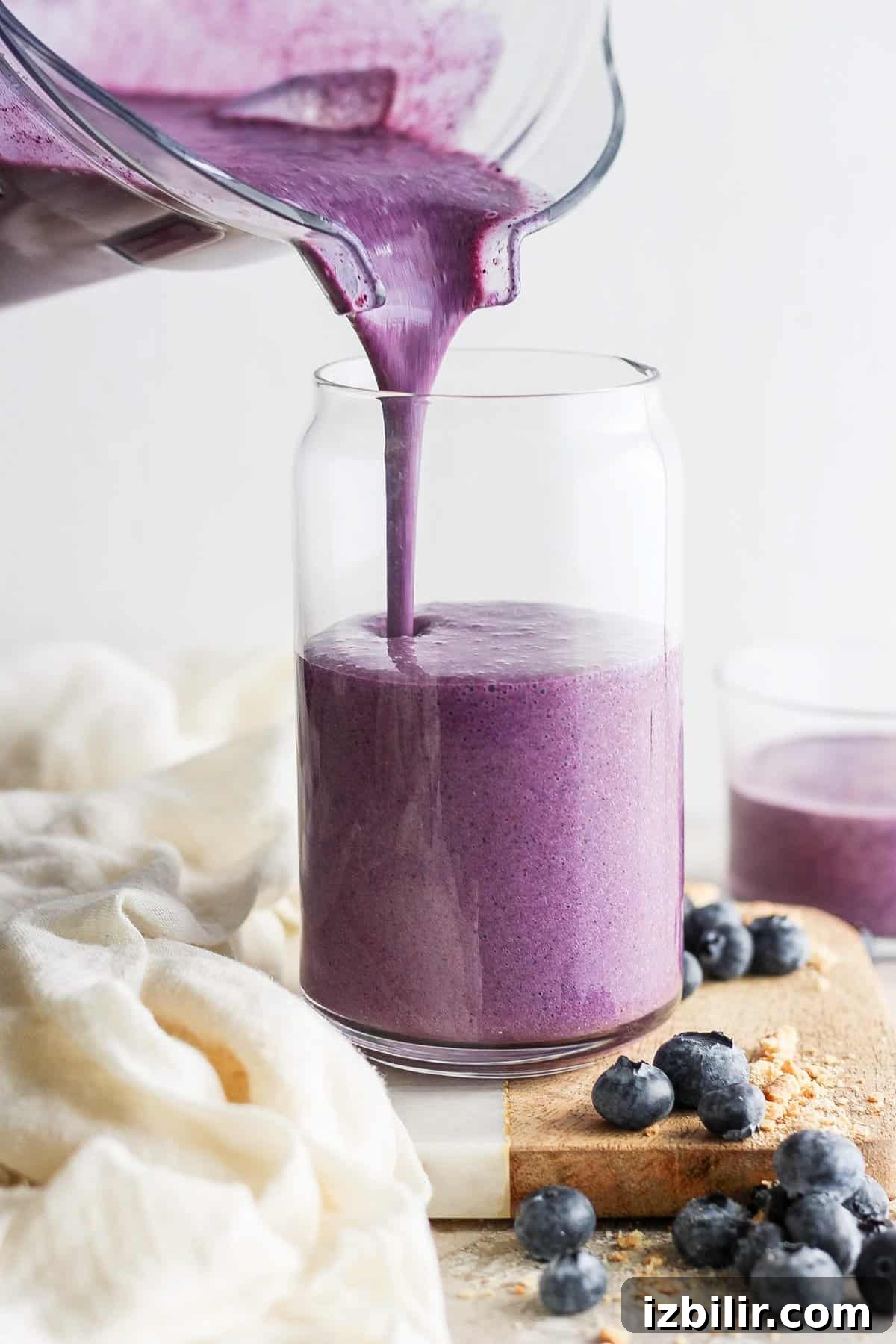 Pouring a fresh blueberry smoothie into a glass jar, surrounded by scattered blueberries on a wooden board.