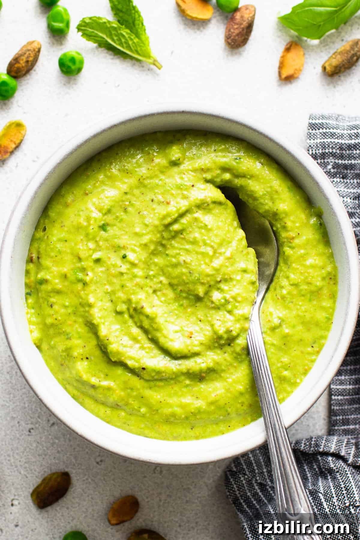 A bowl of creamy green pea and mint soup with a spoon, surrounded by peas, mint leaves, and spices on a light surface.