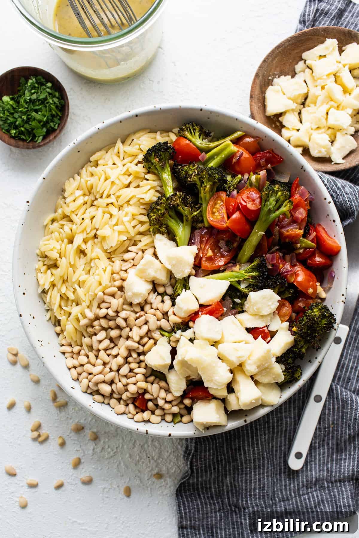 A bowl of salad with orzo pasta, roasted broccoli, cherry tomatoes, mozzarella cheese, and pine nuts on a white surface.