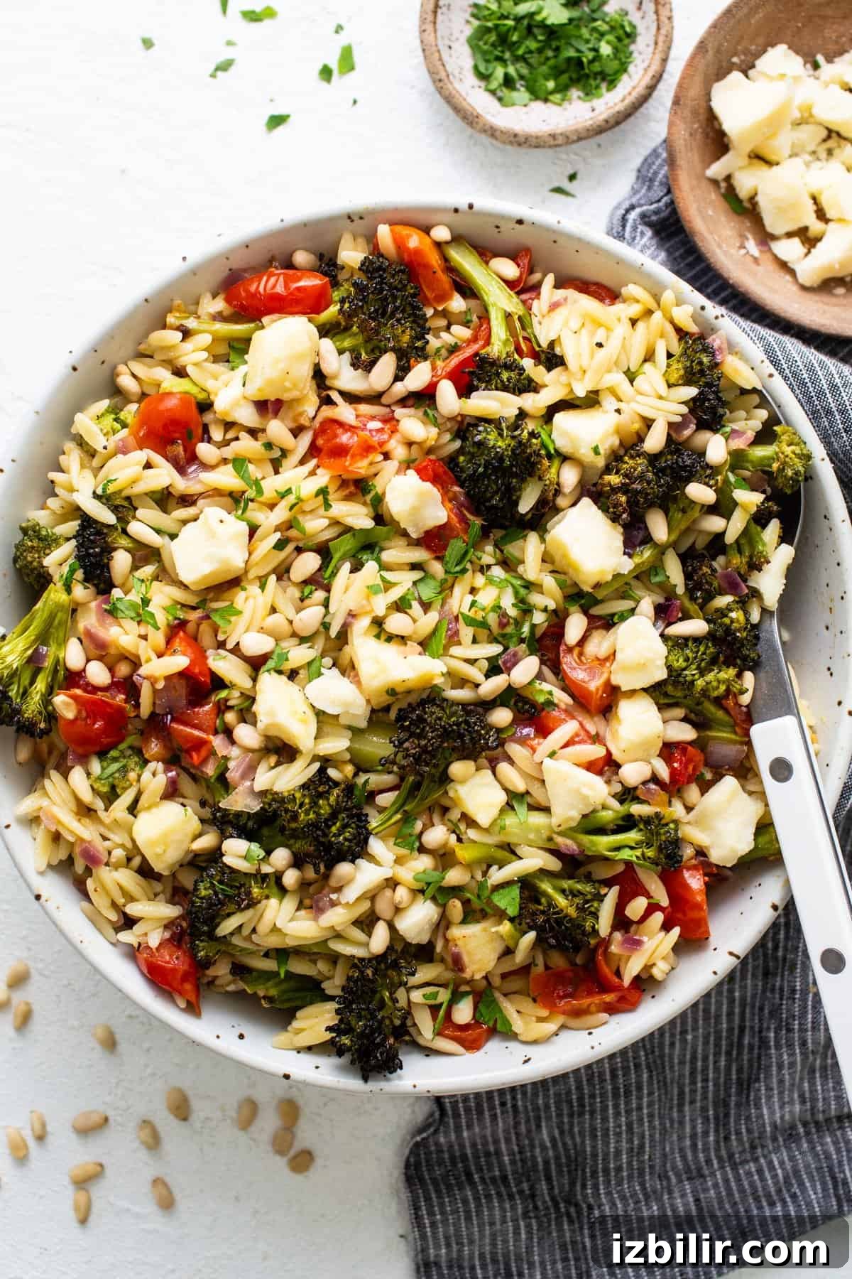 A vibrant orzo salad with broccoli, tomatoes, and pine nuts in a white bowl, garnished with herbs and cheese, on a textured white surface.