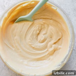 A glass bowl containing creamy peanut butter with a green spoon, photographed from overhead on a textured white surface.