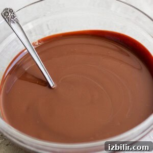 A glass bowl containing smooth melted chocolate with a metal spoon resting inside, set on a light-colored surface.