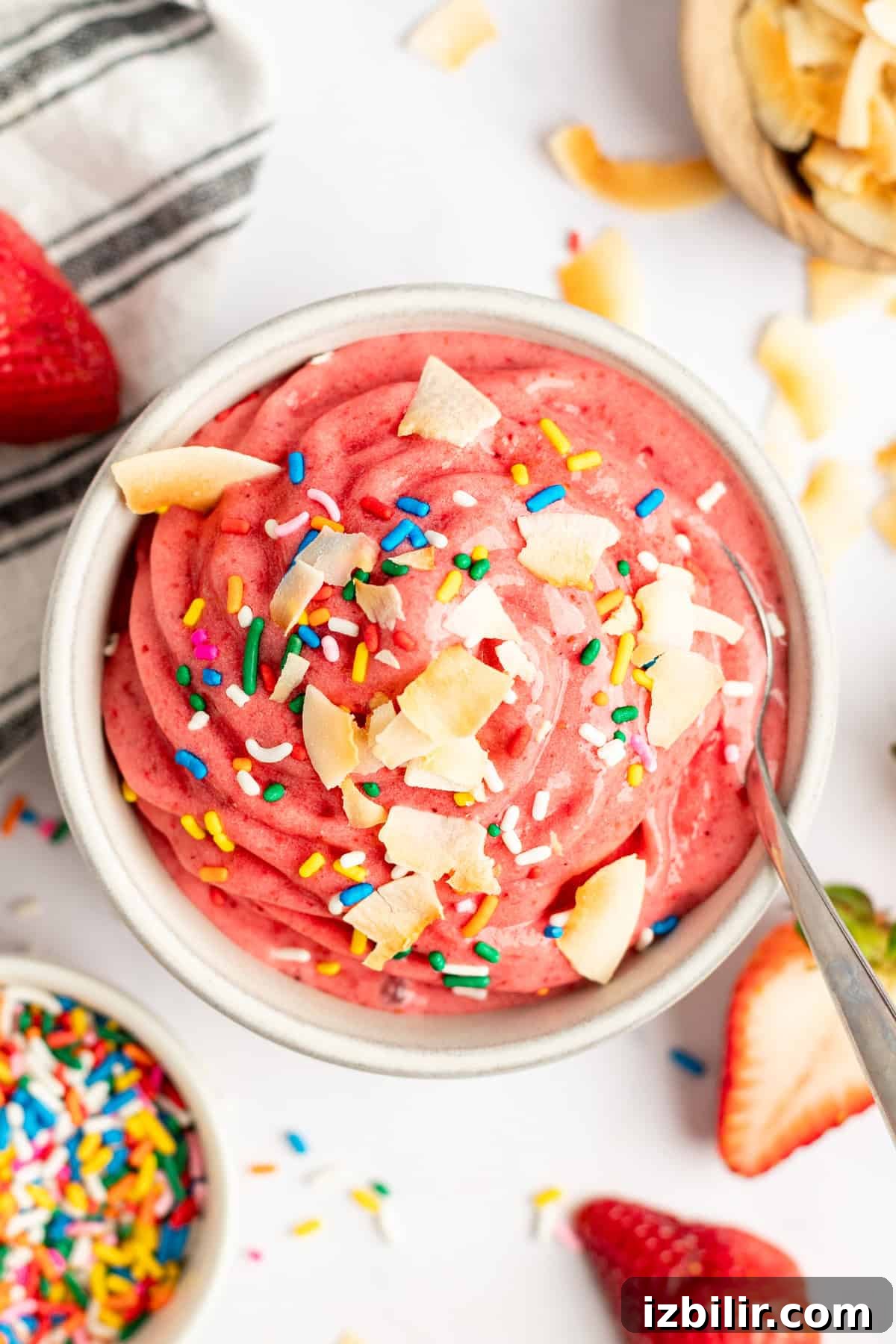 How to Make Strawberry Dole Whip: Dairy Free Soft Serve 2 A vibrant pink smoothie bowl topped with coconut flakes and rainbow sprinkles, accompanied by fresh strawberries and a spoon.