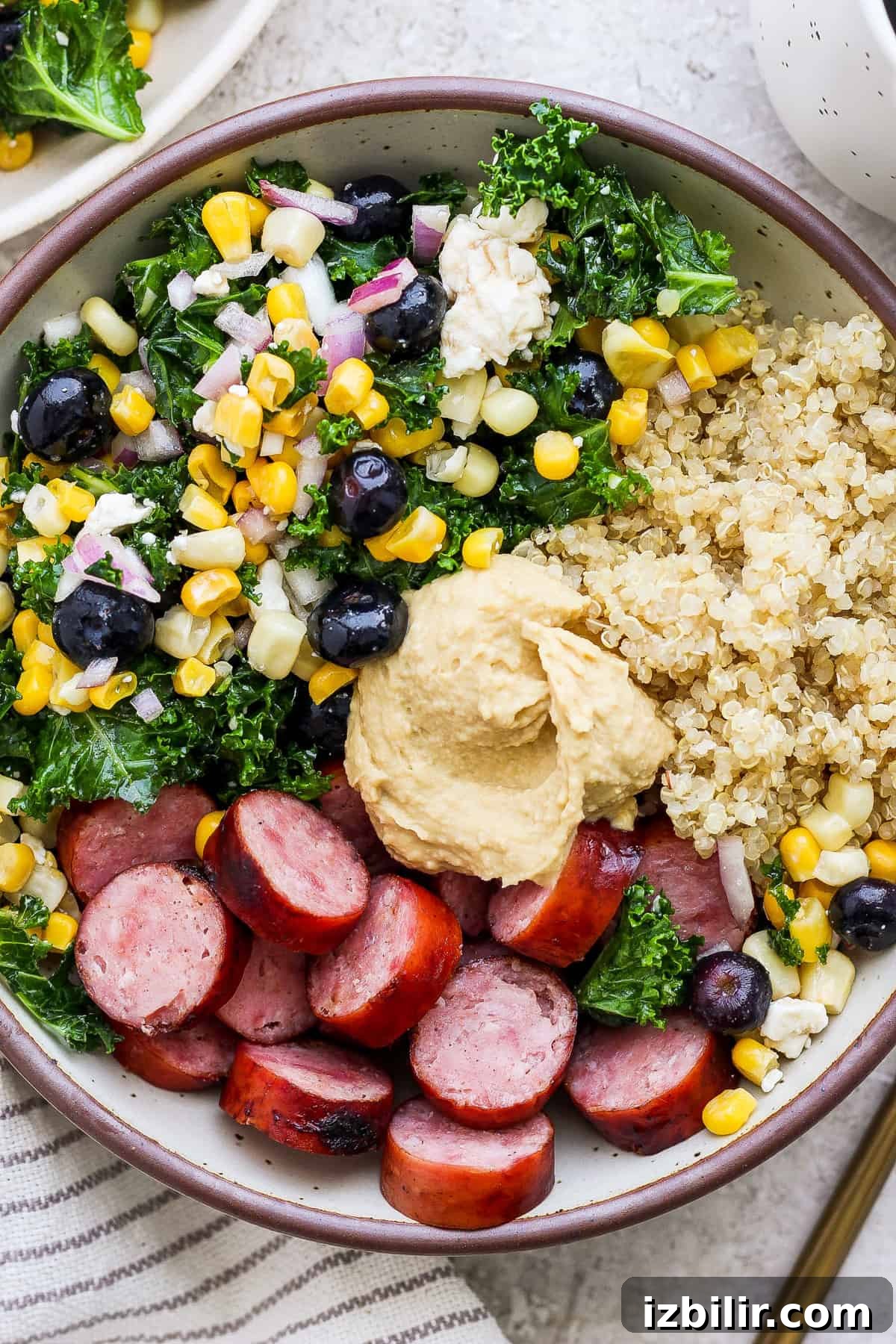 Savory Mixed Protein Bowl with Grains and Veggies 2 A bowl filled with quinoa, sliced sausage, hummus, kale salad, and a mix of corn and blueberries.