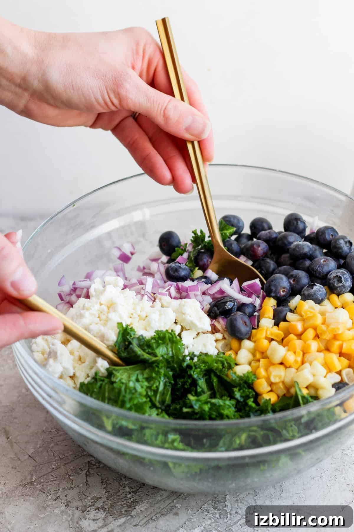 Savory Mixed Protein Bowl with Grains and Veggies 4 A person mixing a colorful salad with blueberries, corn, onions, and greens in a glass bowl using a pair of golden tongs.