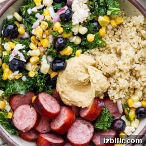 A bowl filled with quinoa, sliced sausage, kale, corn, black olives, onions, and a dollop of hummus.