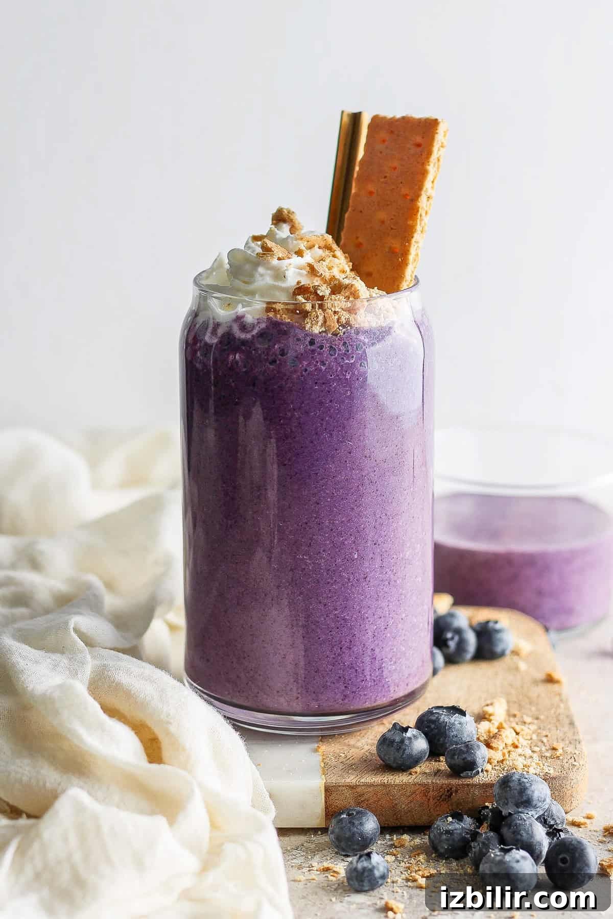 Fit Foodie Cooking Club: May 2024 Seasonal Recipes 5 A vibrant purple smoothie topped with whipped cream and a cookie, garnished with blueberries on a light, textured background.