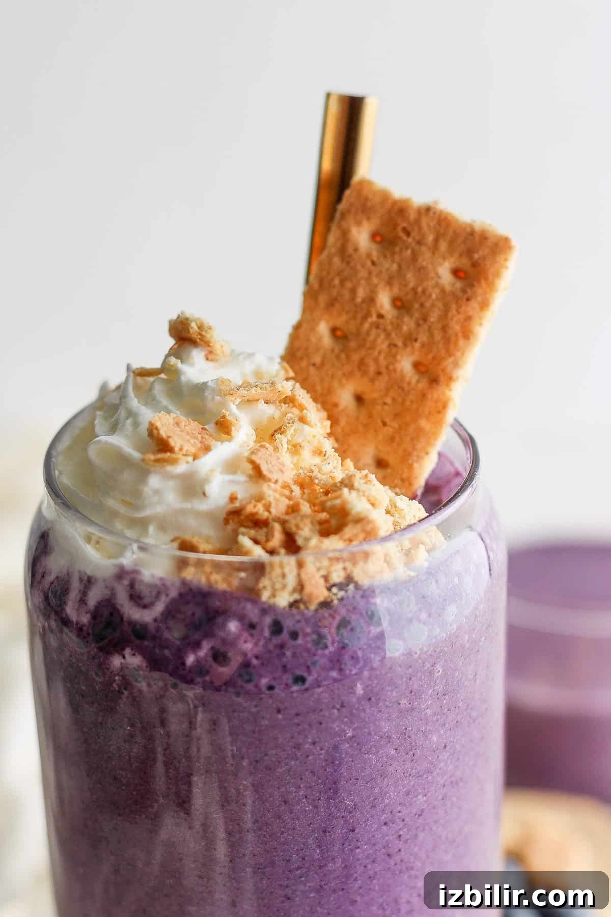 Fit Foodie Cooking Club: May 2024 Seasonal Recipes 6 A vibrant purple smoothie topped with whipped cream and crushed biscuits, served in a clear glass with a gold straw.