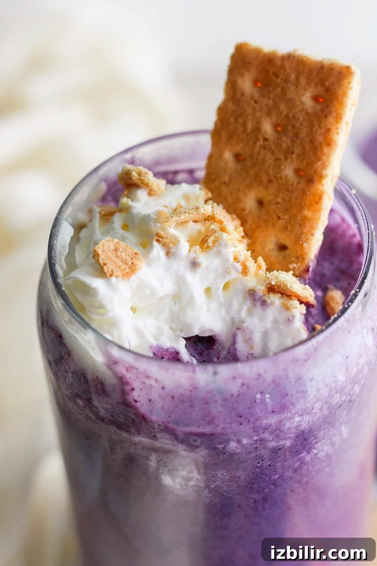 Fit Foodie Cooking Club: May 2024 Seasonal Recipes 7 A close-up of a purple milkshake topped with whipped cream, crumbled cookies, and a cookie sticking out, served in a clear glass.