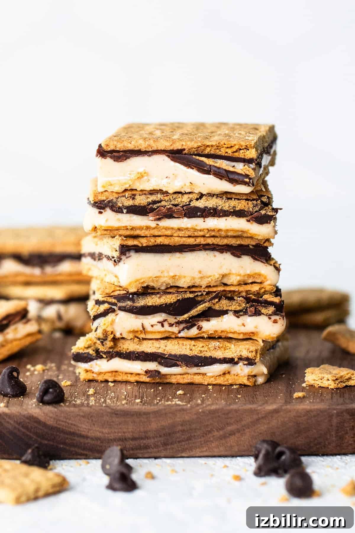 A stack of s'mores bars with layers of graham crackers, melted marshmallow, and chocolate on a wooden board, with scattered chocolate chips around.
