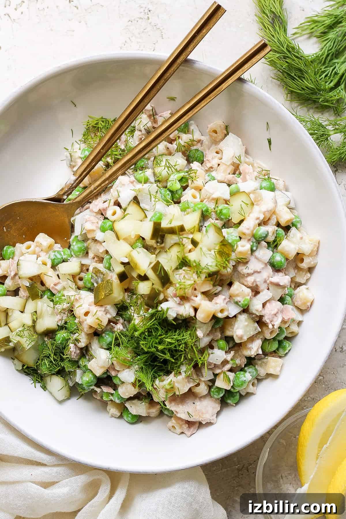Creamy Dill Tuna Pasta Salad Recipe 2 A bowl of tuna salad with peas, chopped cucumber, and dill, served with a pair of chopsticks and fresh lemon slices on a neutral-toned background.