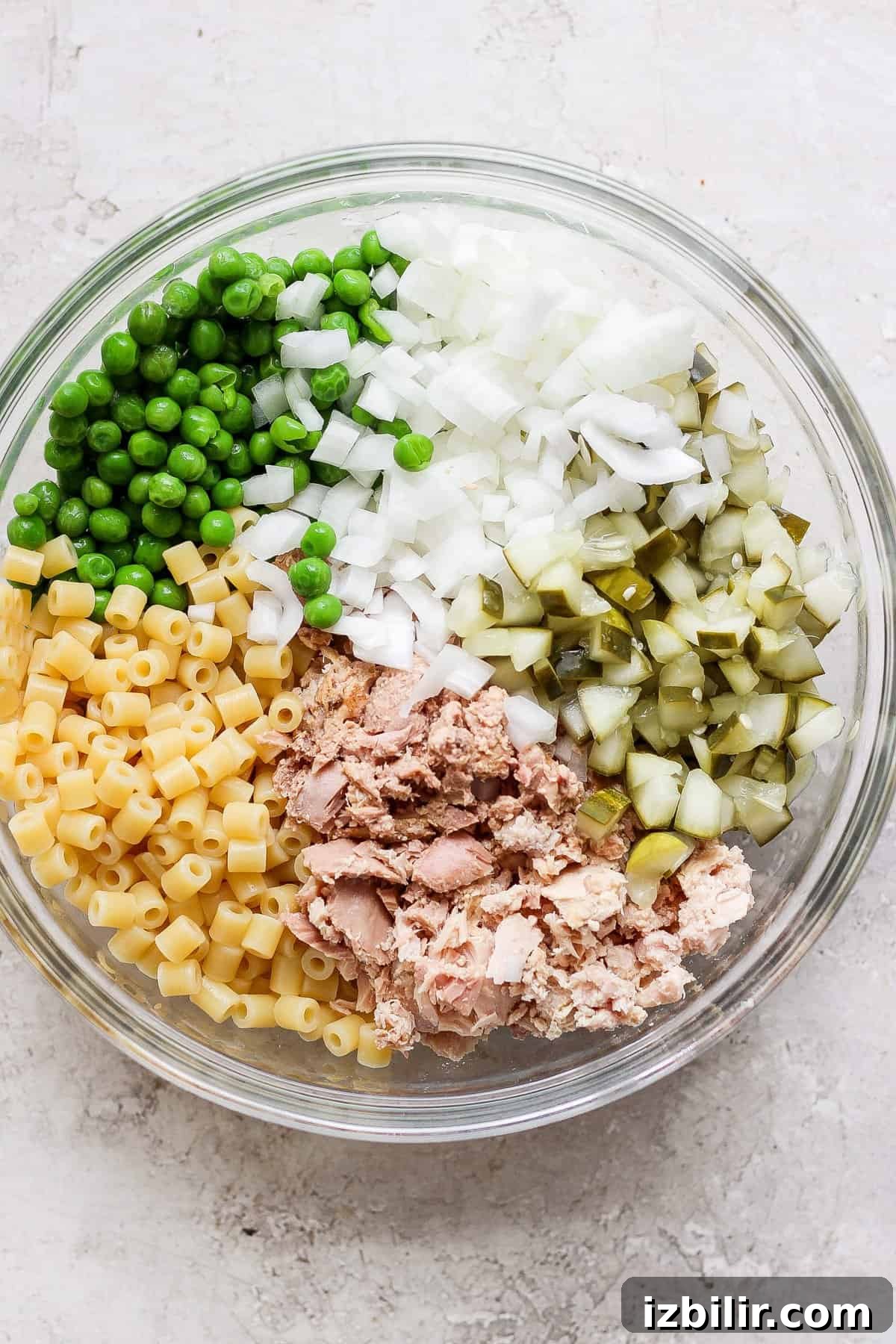Creamy Dill Tuna Pasta Salad Recipe 3 A bowl containing separate sections of diced onions, green peas, chopped pickles, macaroni pasta, and canned tuna, arranged on a light, textured surface.