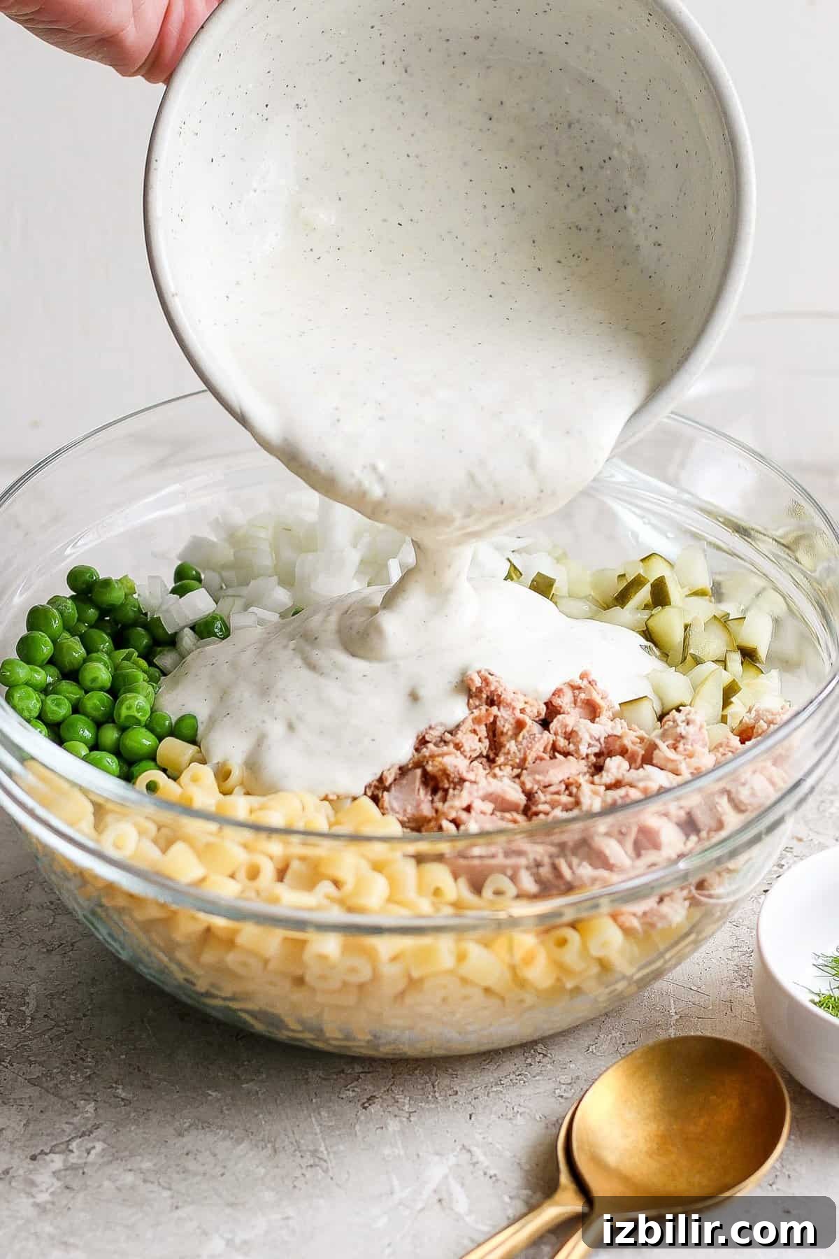 Creamy Dill Tuna Pasta Salad Recipe 4 A person pours creamy sauce into a bowl containing macaroni, tuna, chopped onions, and peas for a salad.