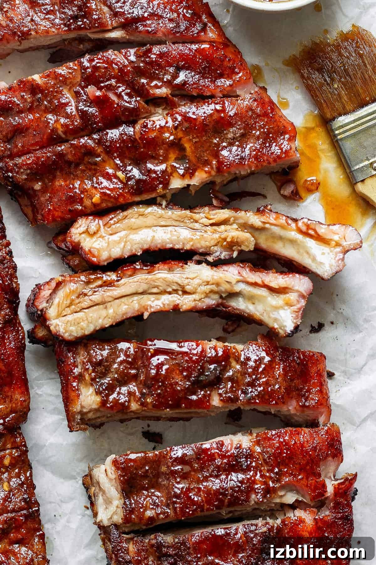 Close-up of cooked, glazed pork ribs on parchment paper with sauce and a basting brush beside them.