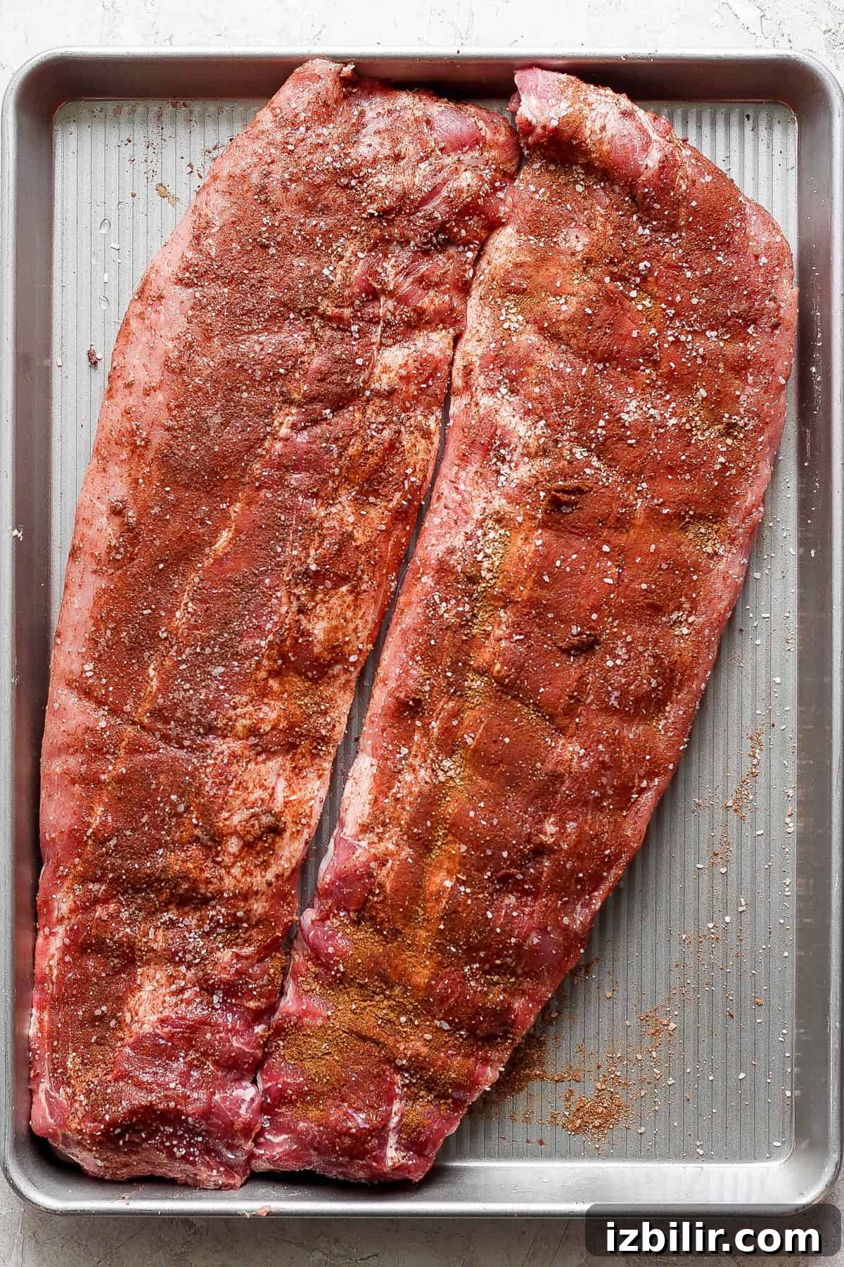 Two slabs of seasoned raw pork ribs are placed on a large rectangular baking sheet, ready for cooking.