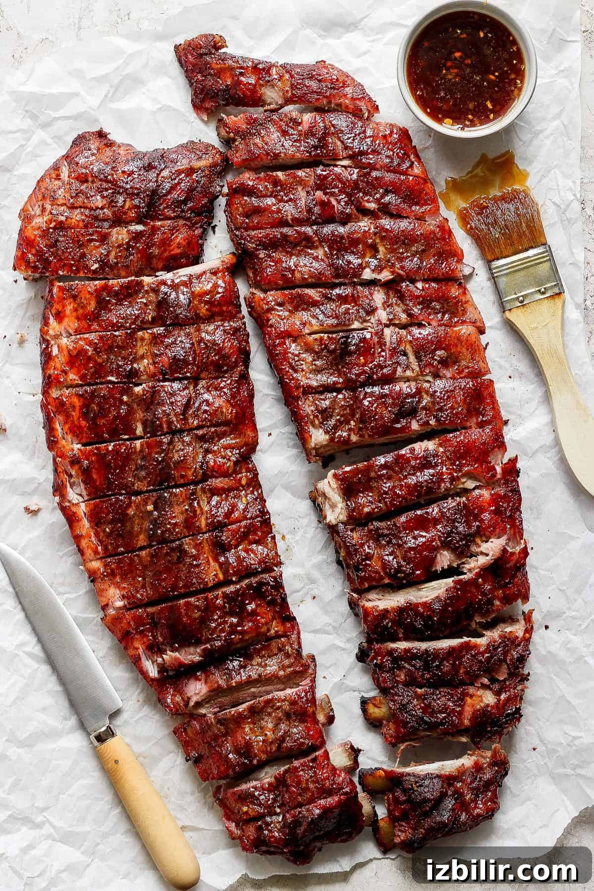 Two racks of barbecued ribs cut into portions, placed on parchment paper with a knife, a brush, and a bowl of sauce.