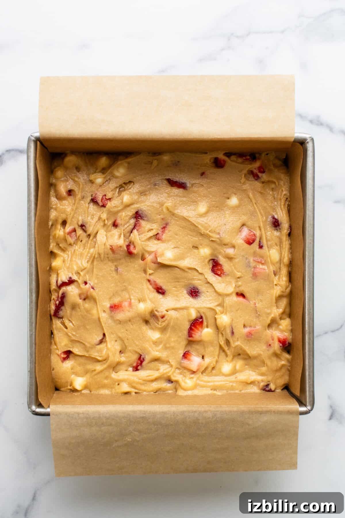 White Chocolate Strawberry Blondies Recipe 3 Square baking pan lined with parchment paper and filled with raw batter containing chunks of fruit, promising delicious strawberry blondies on a marble countertop.