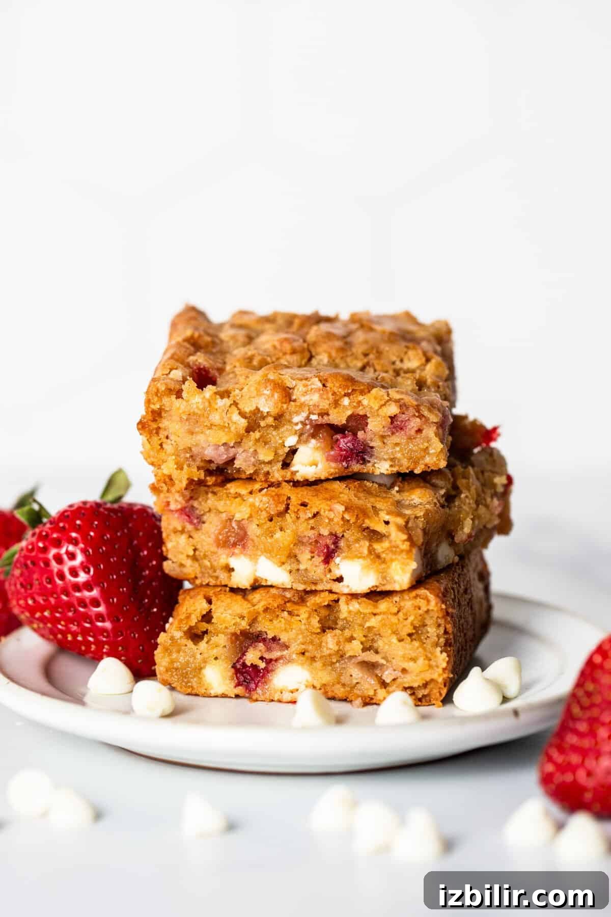 White Chocolate Strawberry Blondies Recipe 5 A plate of three stacked strawberry blondies containing white chocolate chips and strawberry pieces, surrounded by strawberries and white chocolate chips.