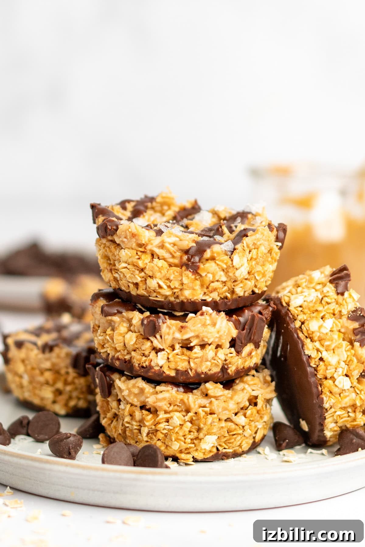 A stack of four oat and chocolate dessert bars on a plate with chocolate chips scattered around. A jar with peanut butter is visible in the background.