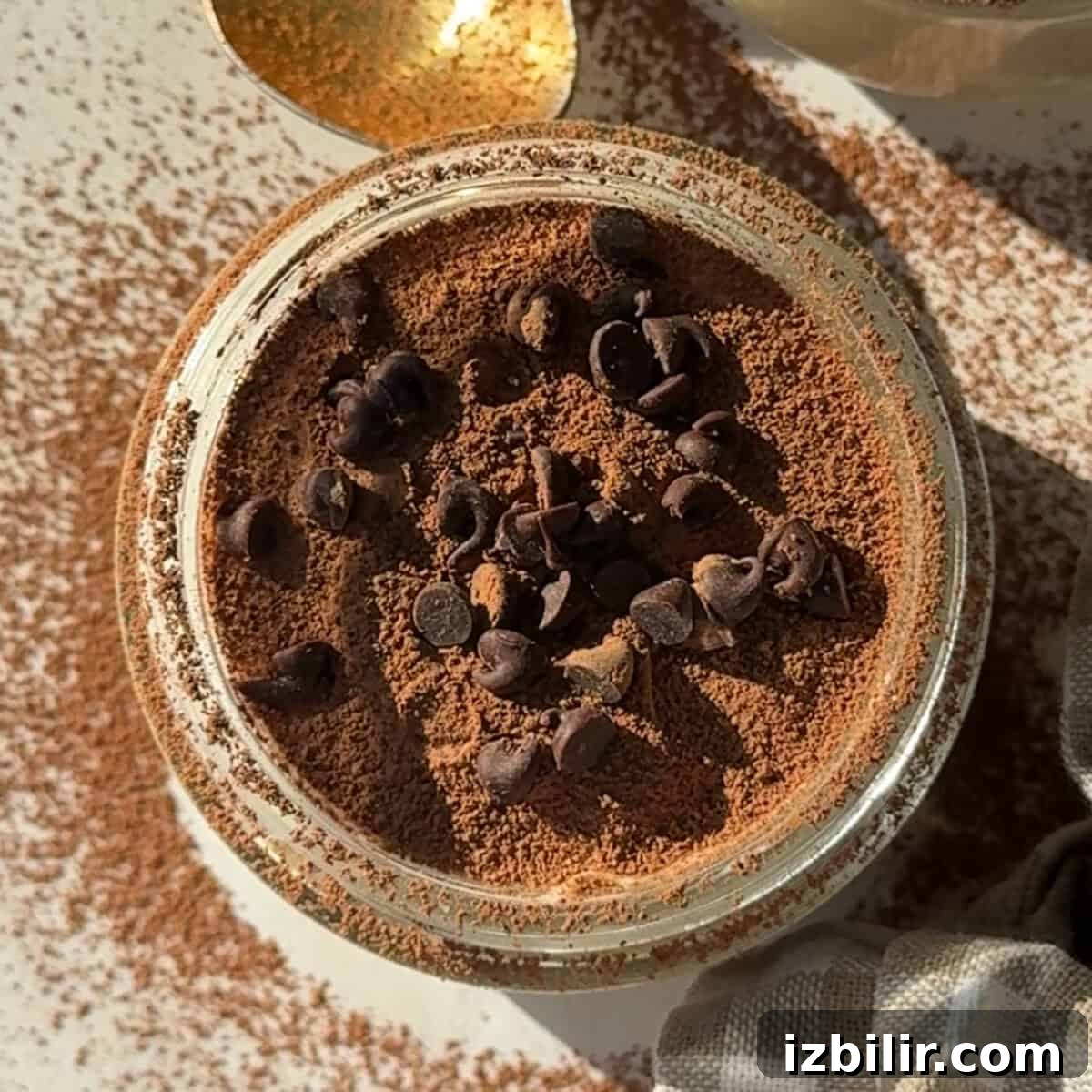 A jar filled with a powdered brown substance topped with chocolate chips. A gold spoon is partially visible nearby, with more powder scattered on the surface.