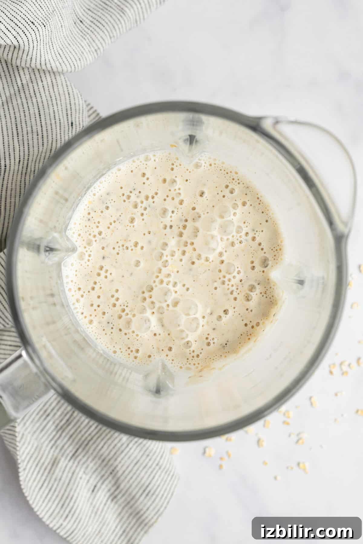 A blender filled with a frothy mixture is shown from above on a light-colored surface. A striped cloth and some spilled oats are visible next to it.