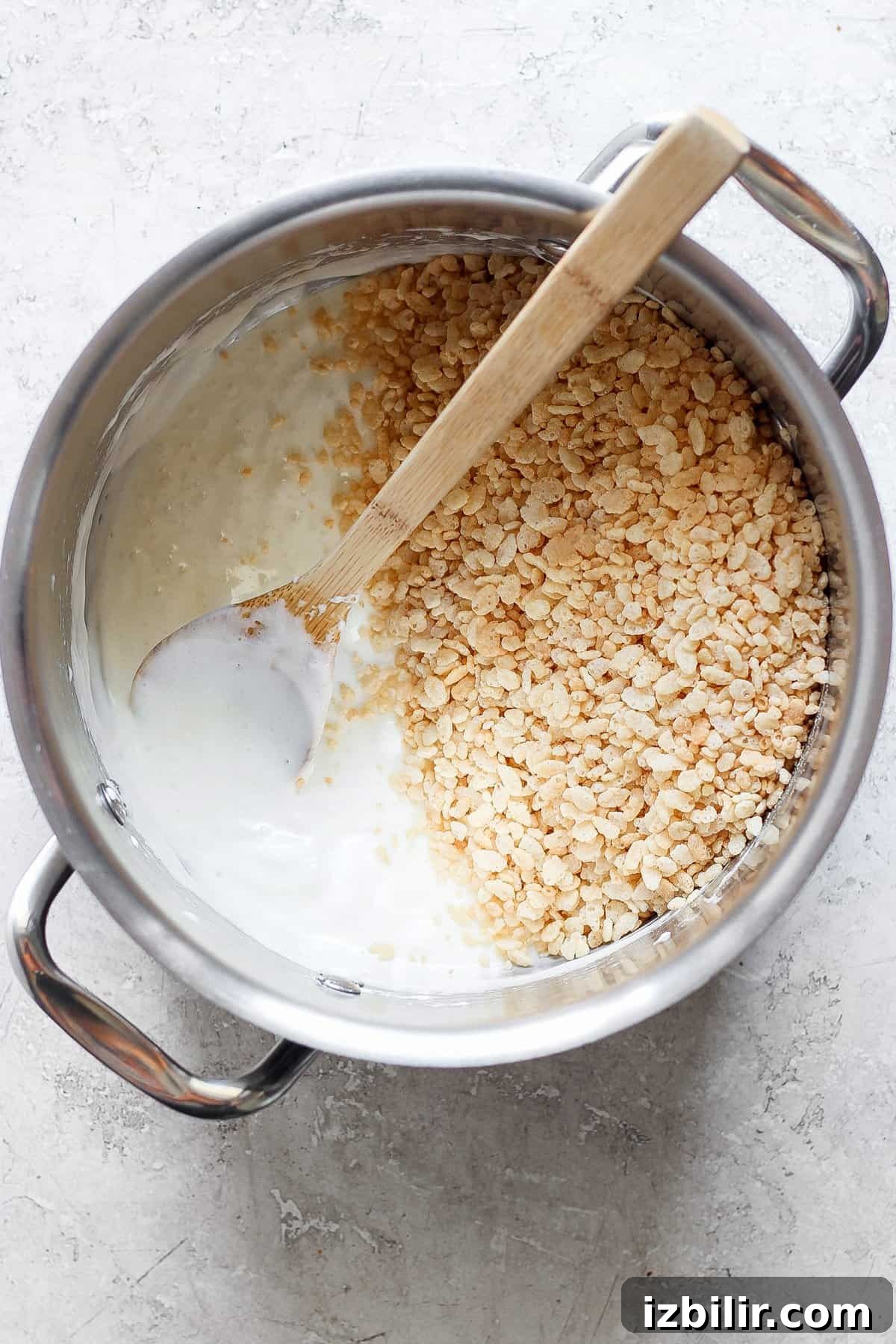 Toasted Marshmallow Rice Krispies Treats Recipe 3 A pot filled with melted marshmallows and some toasted Rice Krispie treats, with a wooden spoon resting inside, on a light-colored surface.