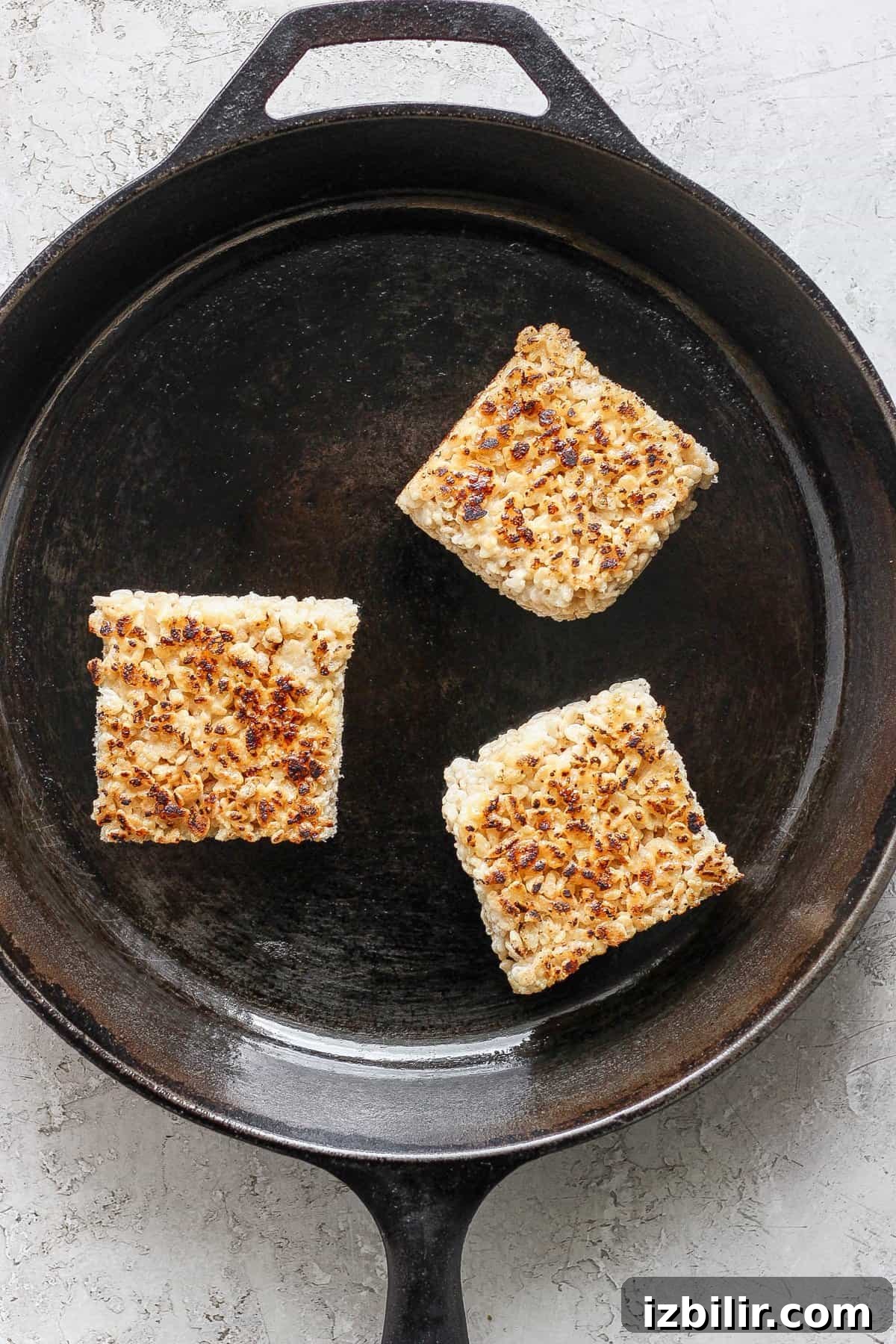 Toasted Marshmallow Rice Krispies Treats Recipe 4 Three toasted Rice Krispie treats in a cast iron skillet on a light textured surface.