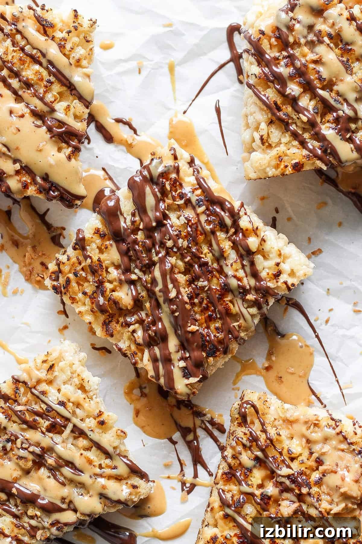 Toasted Marshmallow Rice Krispies Treats Recipe 5 Close-up of five toasted Rice Krispie treats drizzled with chocolate and peanut butter, arranged on parchment paper. The treats are topped with crushed nuts.