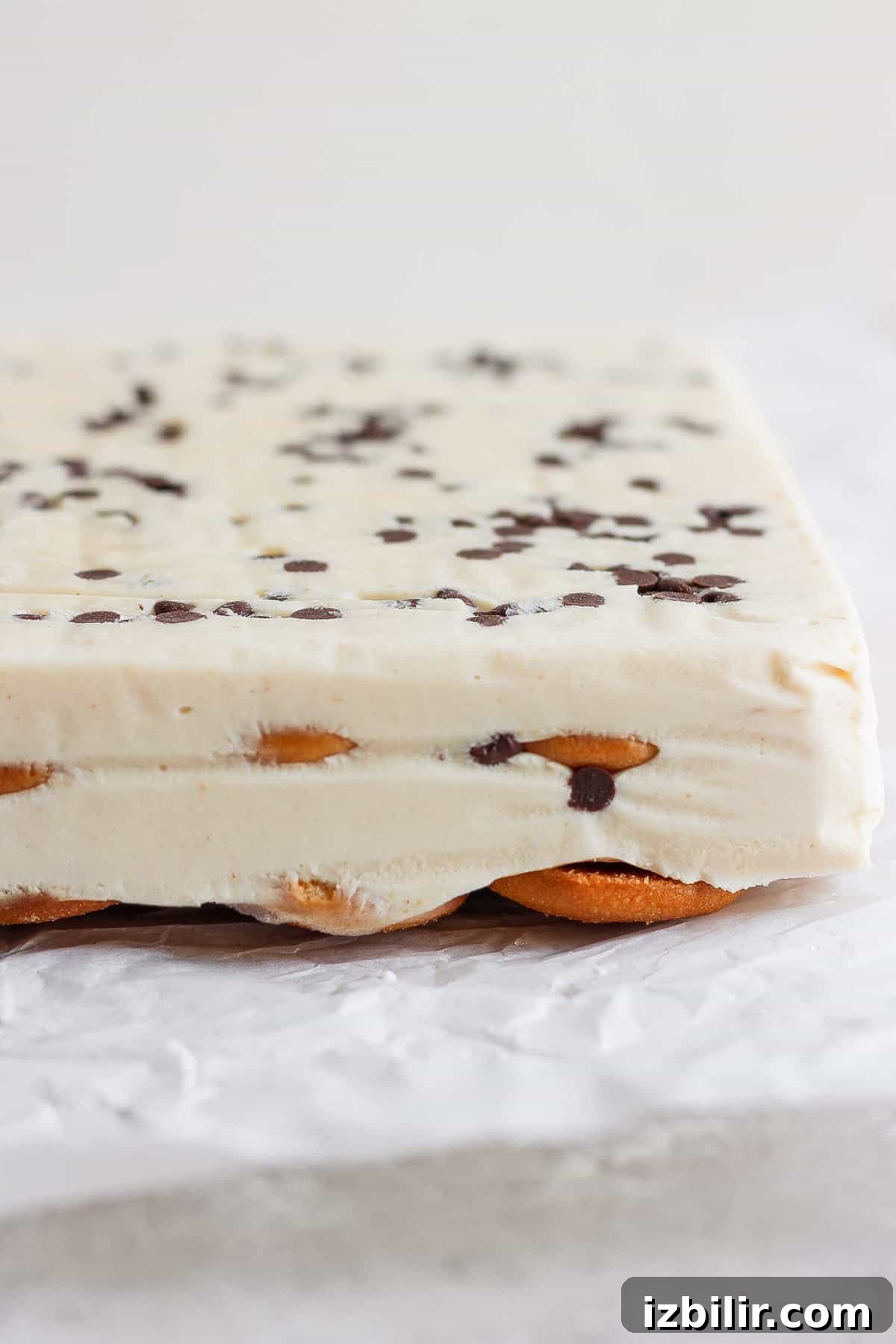 Close-up of a layered peanut butter banana ice box cake with a creamy white filling, chocolate chips on top, and a base of cookies, set on crinkled parchment paper.