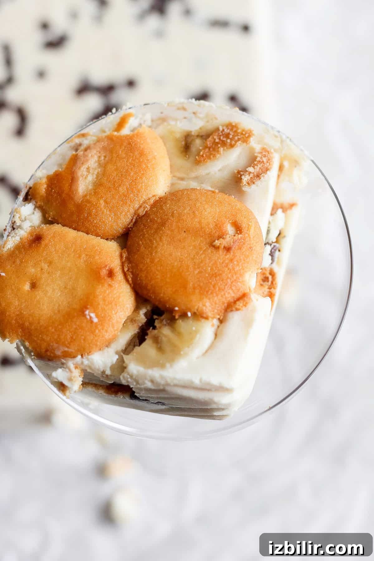 A glass bowl filled with a dessert made of layered vanilla wafers, slices of banana, and creamy pudding. The dessert, reminiscent of a peanut butter banana ice box cake, is topped with vanilla wafers.