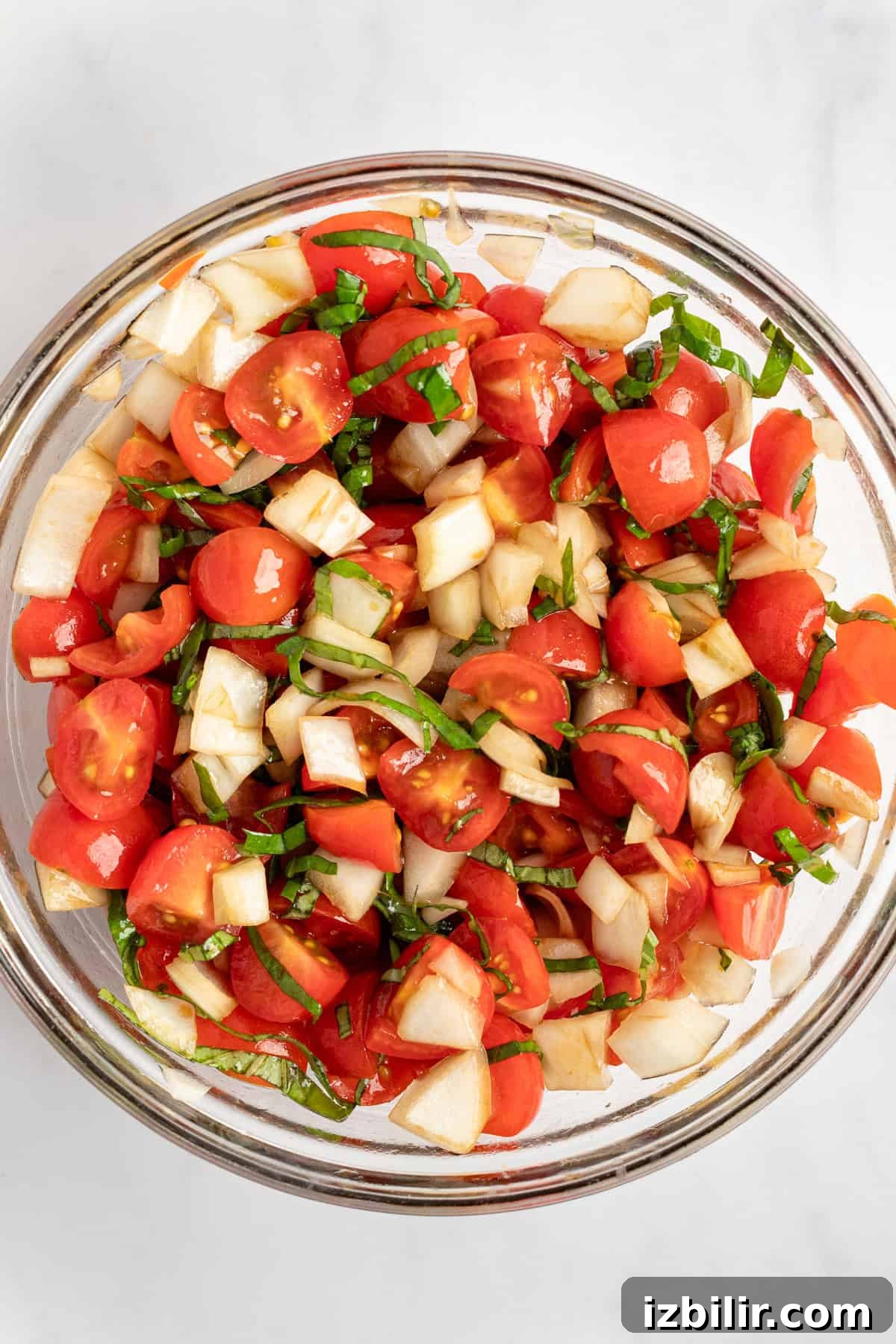 A glass bowl filled with a fresh salad of chopped tomatoes, cucumbers, and herbs on a white surface.