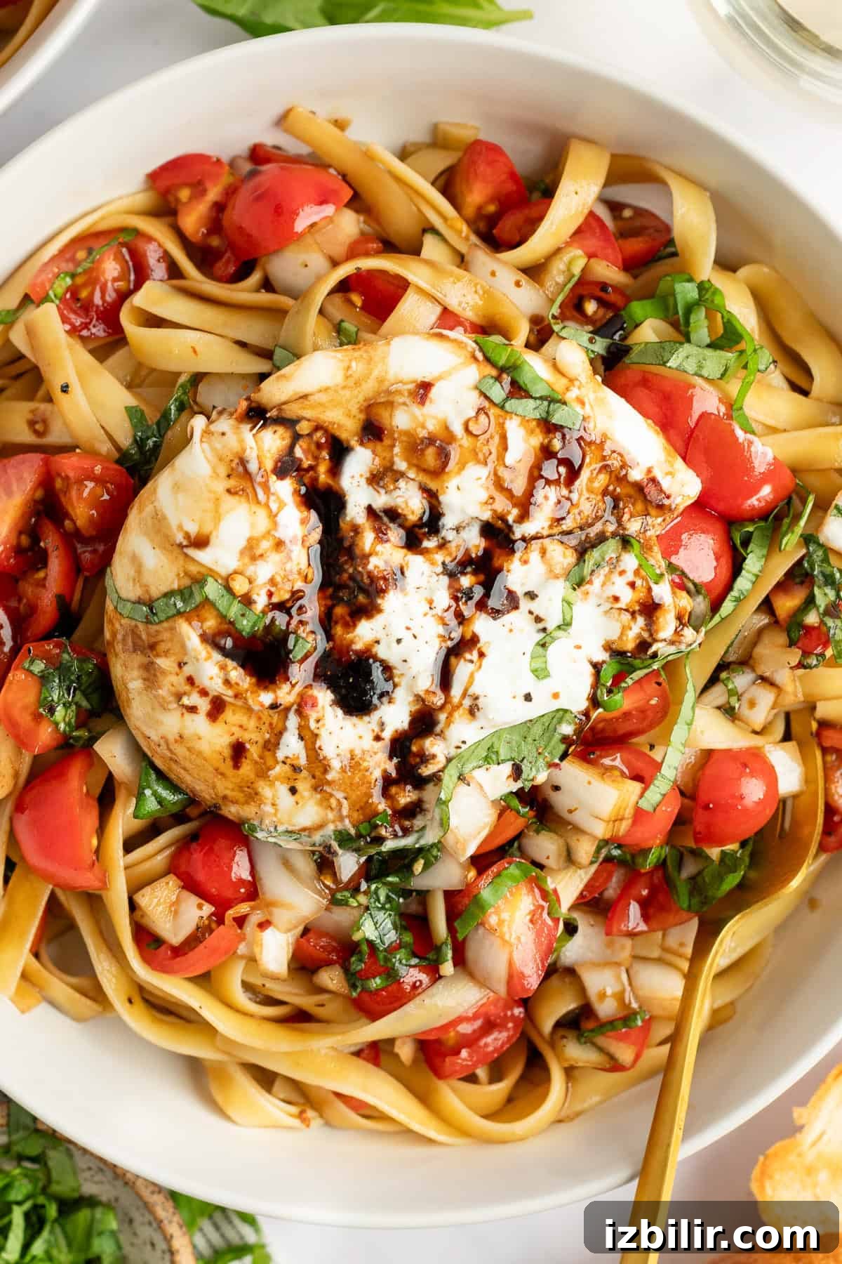 A dish of pasta topped with a large burrata cheese, drizzled with balsamic glaze, and garnished with chopped tomatoes and basil.