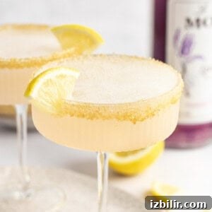 Two cocktails with frothy tops in coupe glasses, each garnished with a lemon wedge and a sugar-rimmed edge. A bottle of purple liquid is partially visible in the background.