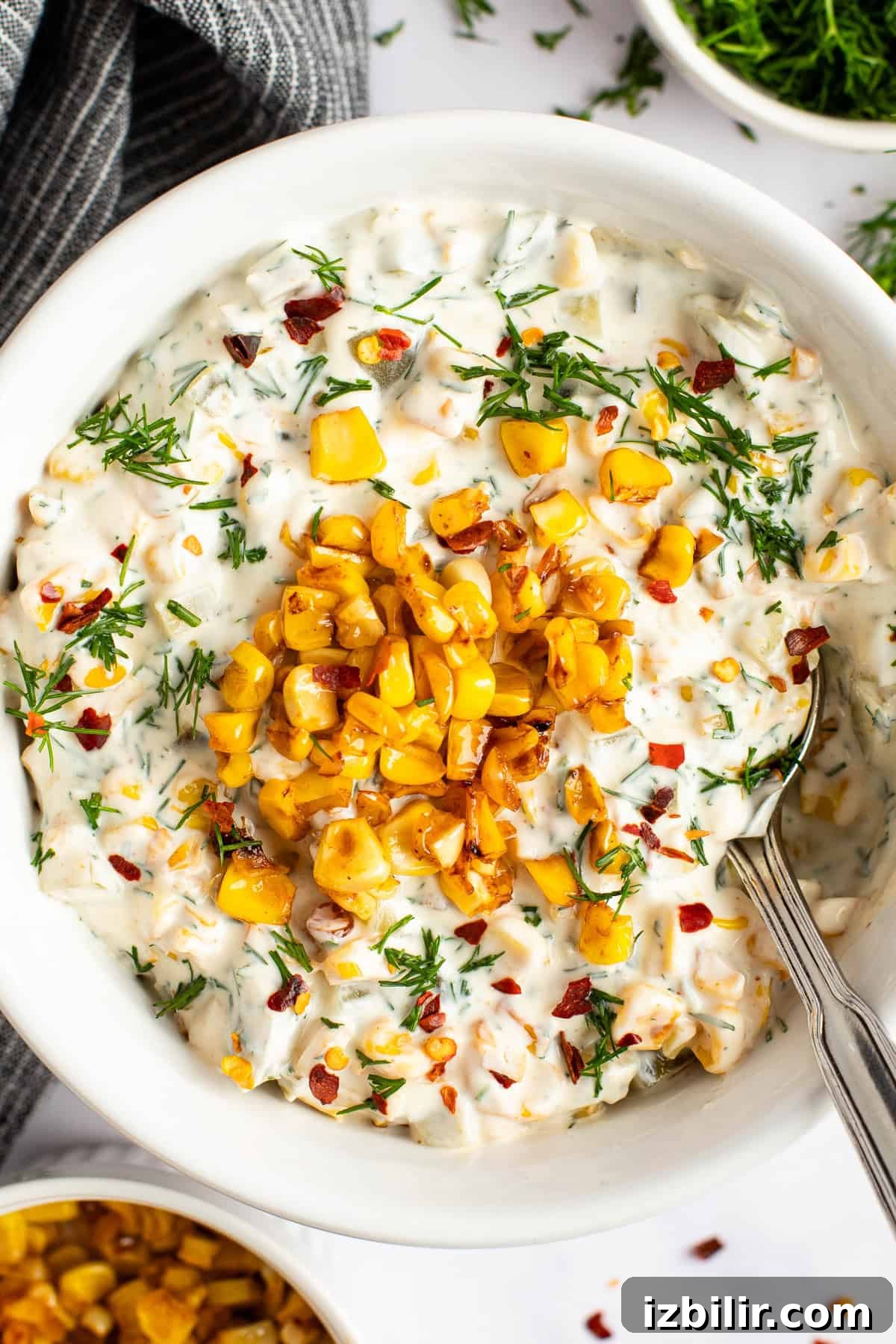A bowl of creamy corn dip garnished with grilled corn, fresh herbs, and red pepper flakes, with a spoon resting inside the bowl.