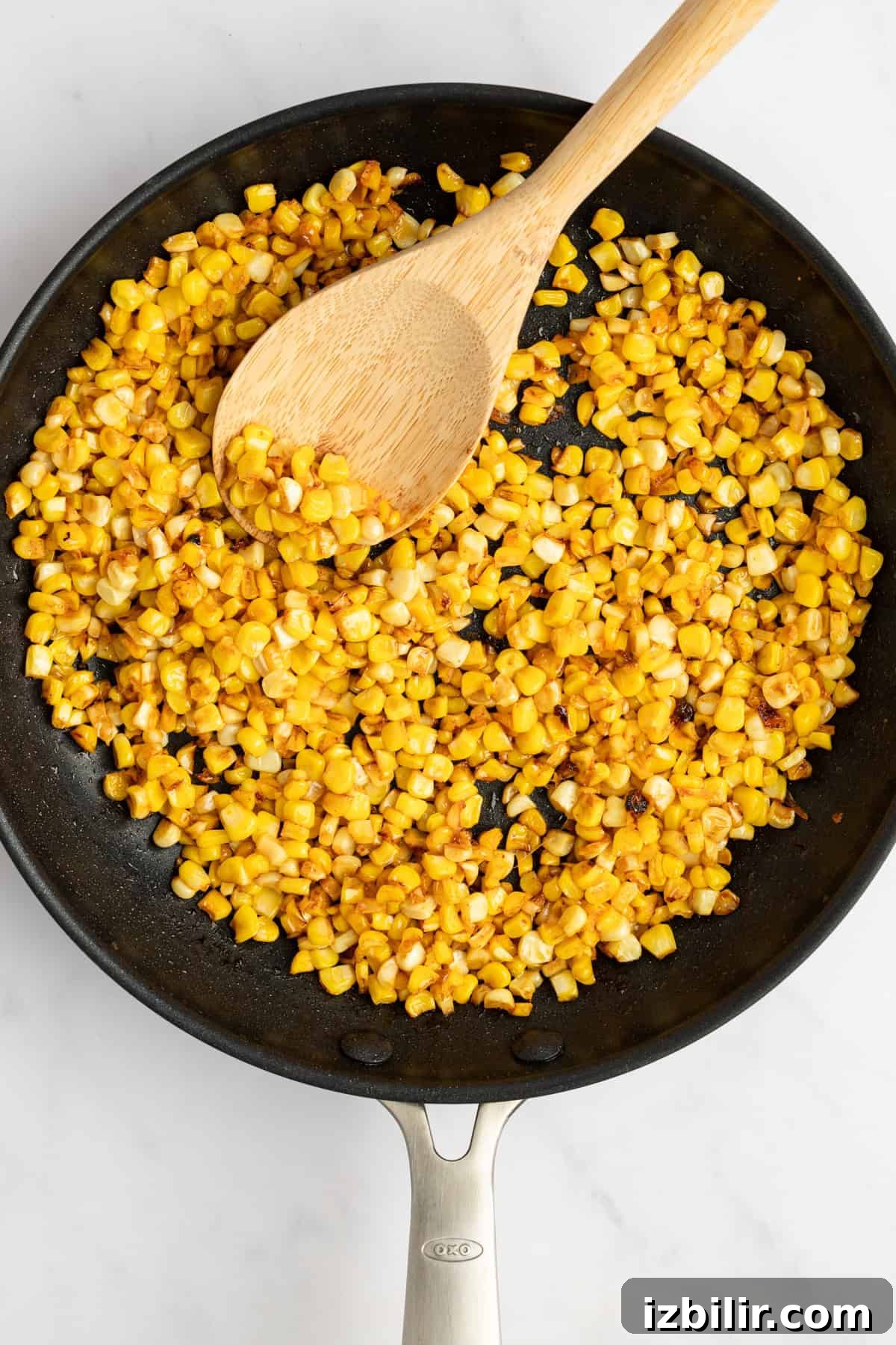 Sautéed corn kernels in a black frying pan with a wooden spoon on a white countertop.