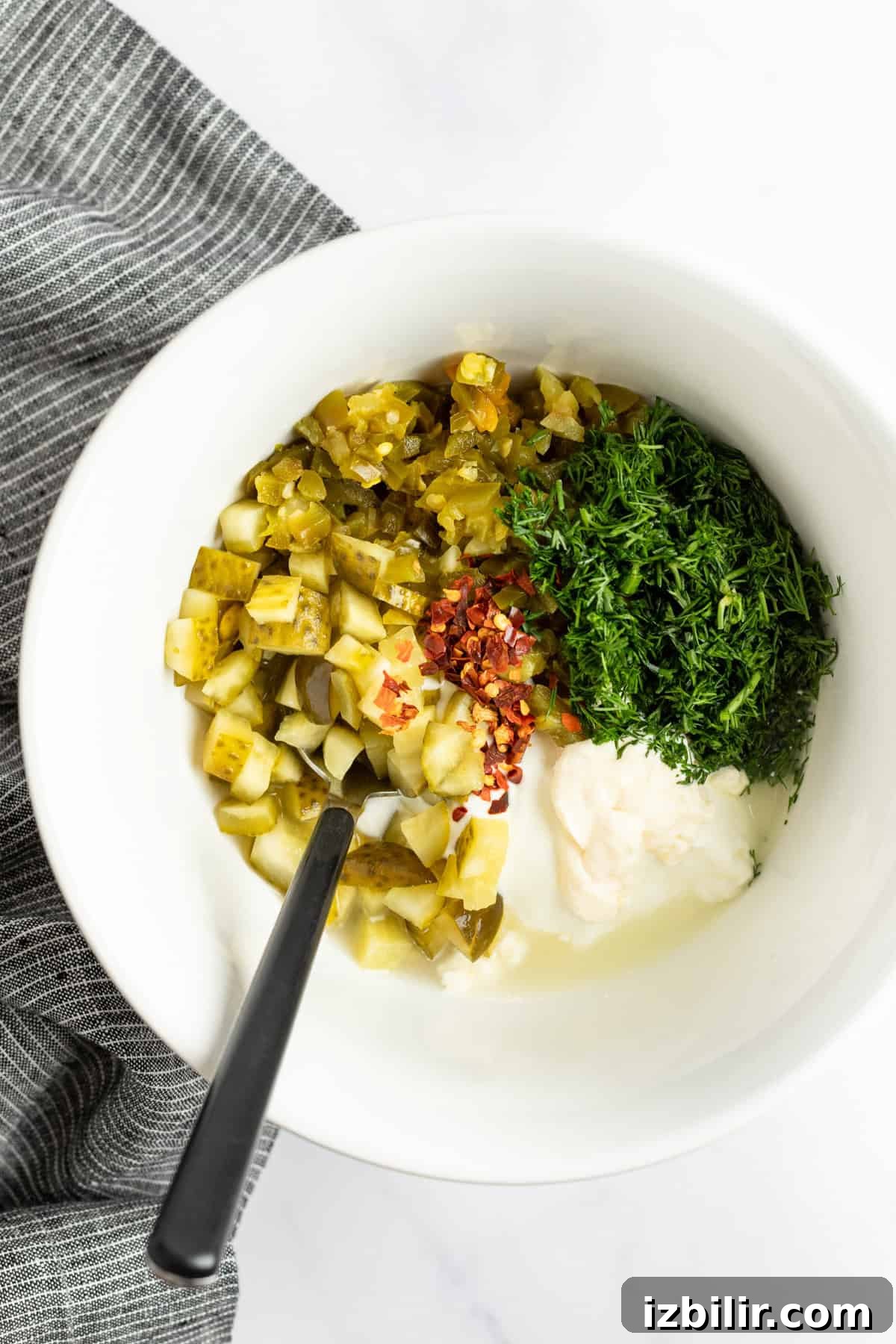 A white bowl holds chopped pickles, fresh herbs, crushed red pepper, and a dollop of creamy white sauce, with a black spoon resting inside. A gray-striped cloth is partially visible on the side.