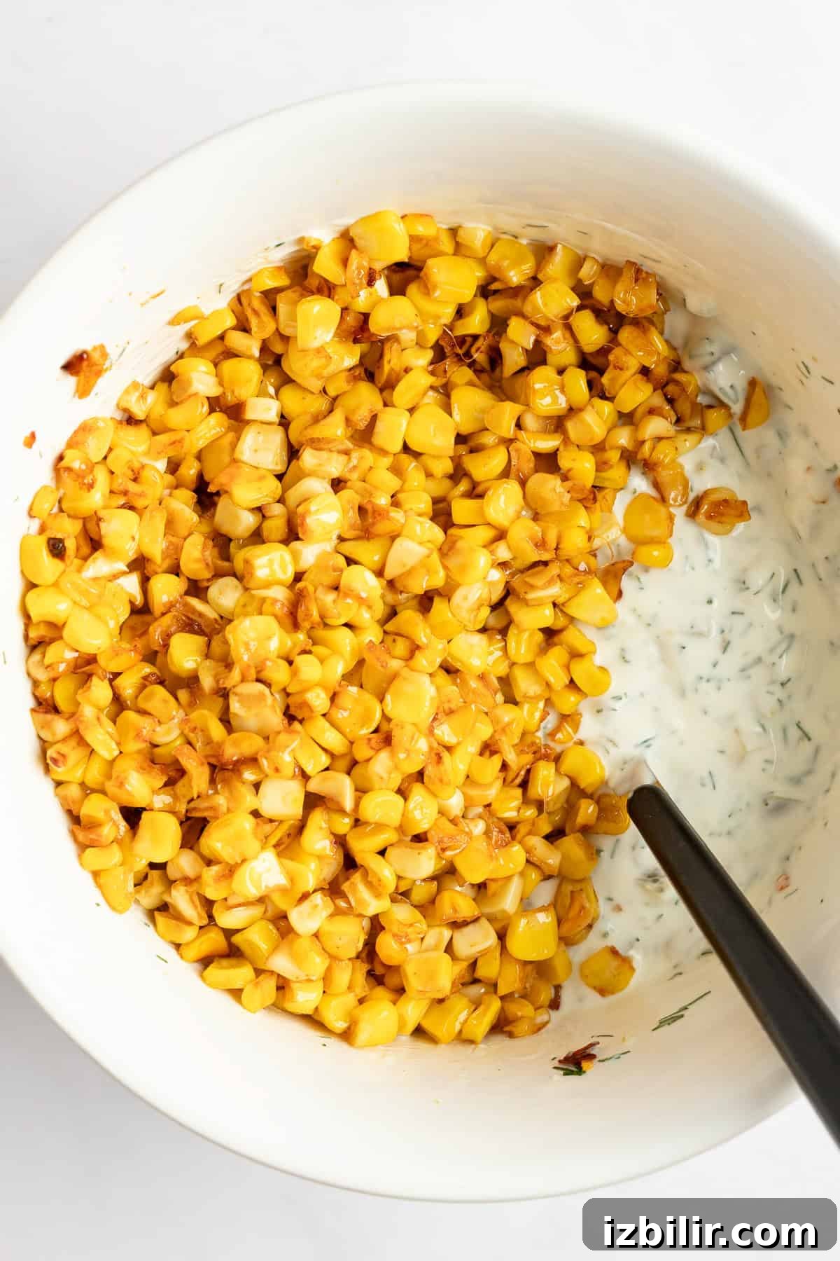 A bowl of roasted corn kernels mixed with a creamy, herb-infused dressing, and spoon partially immersed.