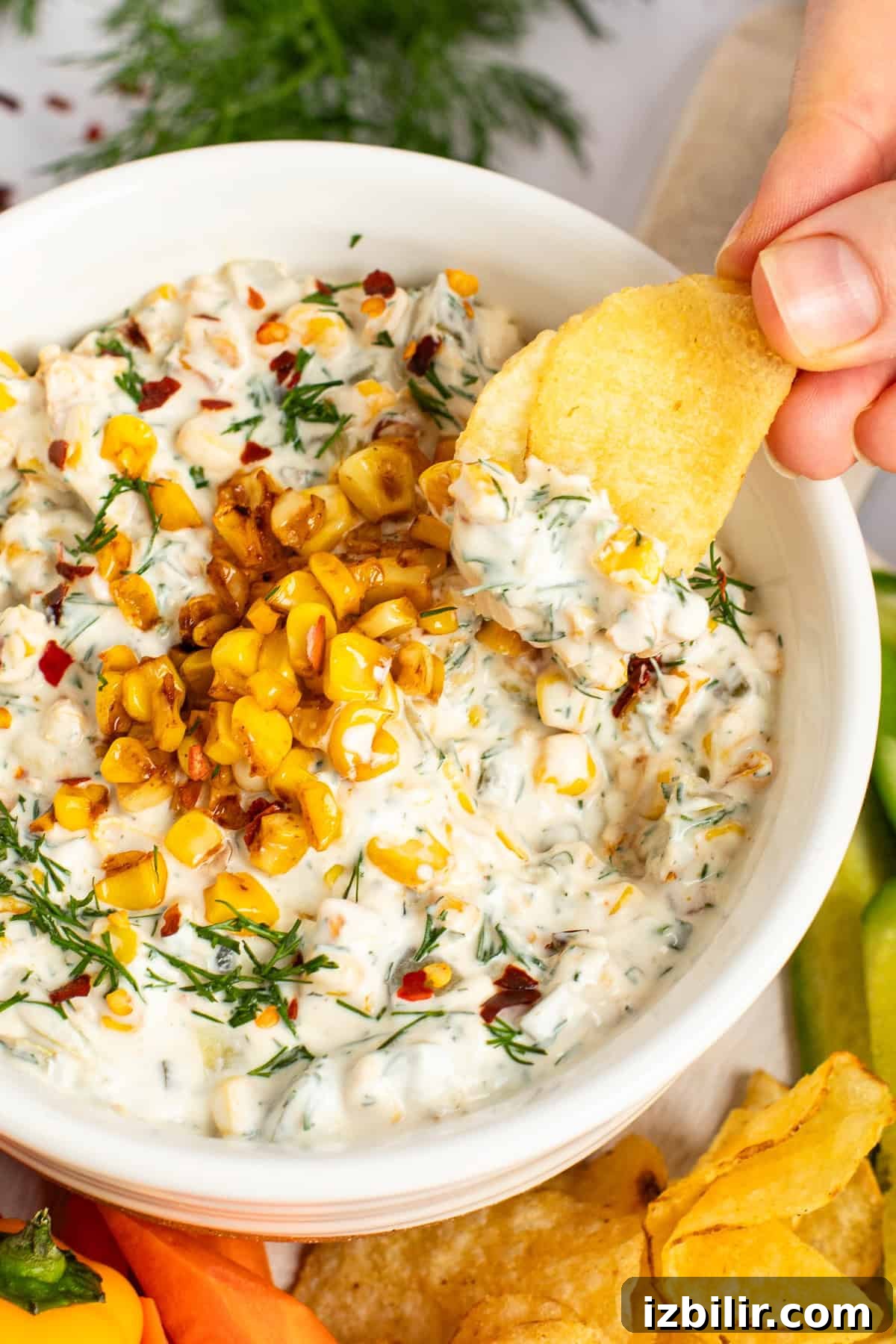 A hand dips a chip into a bowl of creamy dip topped with corn kernels and herbs, surrounded by chips and vegetable slices.