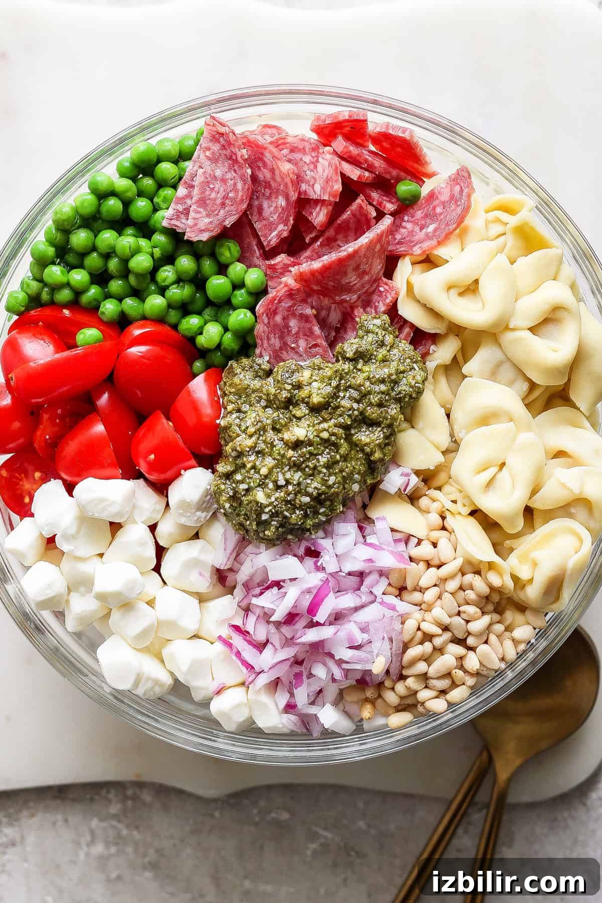 A glass bowl filled with peas, salami slices, cherry tomatoes, mozzarella balls, diced red onions, pine nuts, tortellini, and a dollop of pesto in the center. Two wooden utensils lie beside the bowl.
