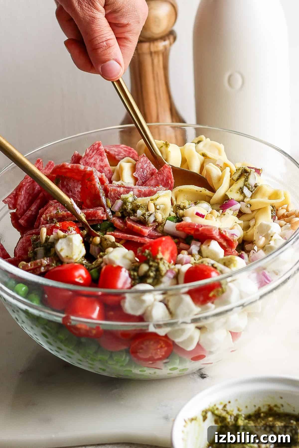 A glass bowl containing ingredients like cherry tomatoes, small mozzarella balls, salami slices, and pasta being mixed with spoons. A bottle and wooden salt grinder are in the background.