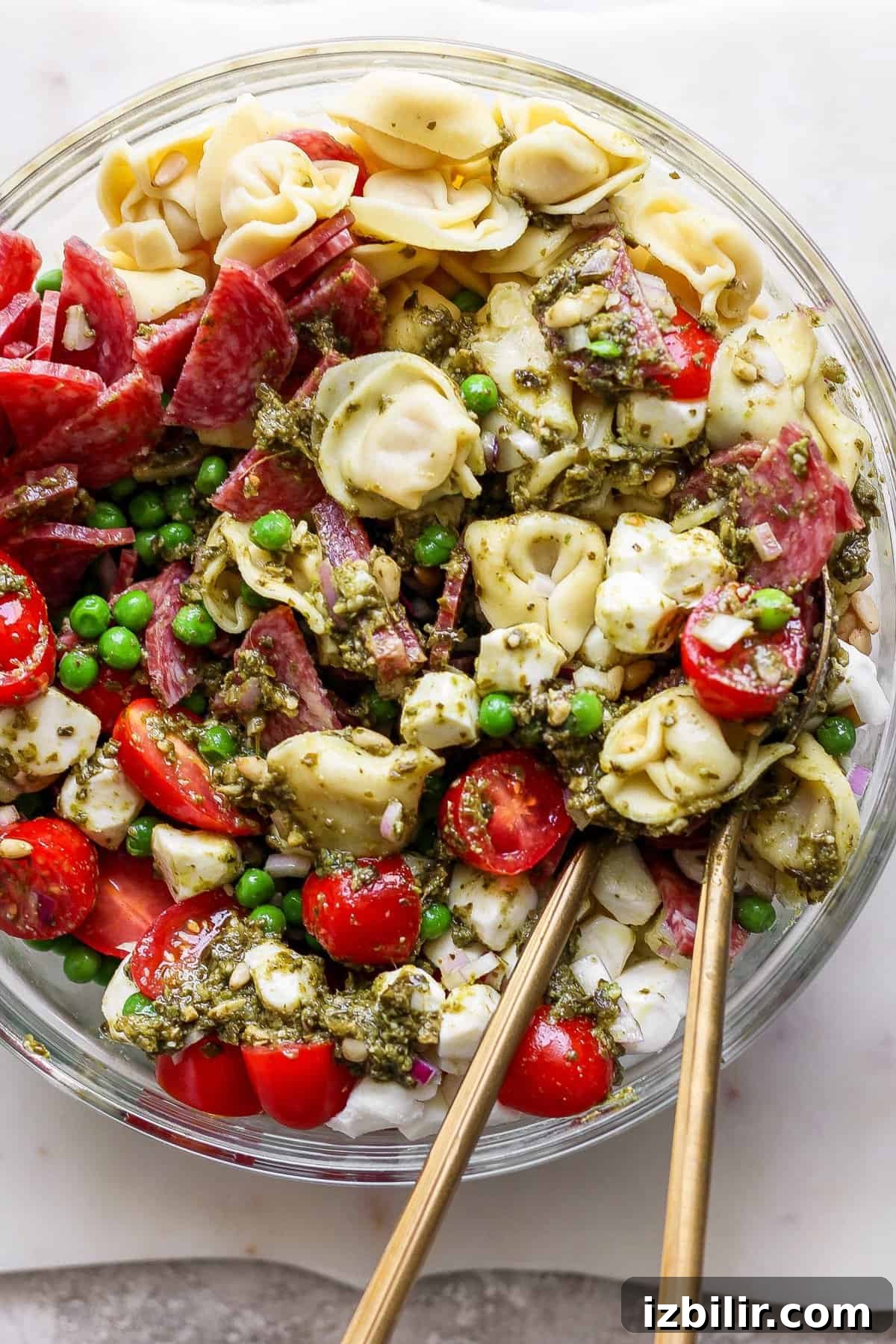 A bowl of pasta salad with tortellini, cherry tomatoes, peas, sliced meat, and pesto sauce, with two wooden utensils placed inside.