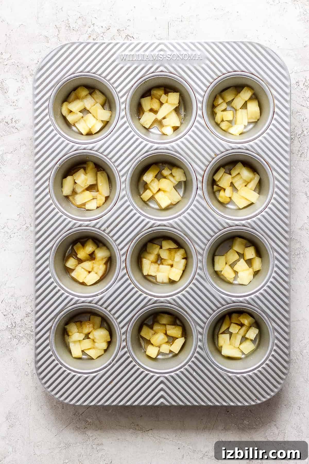 A muffin tin containing pineapple chunks in each compartment, set on a light textured surface.