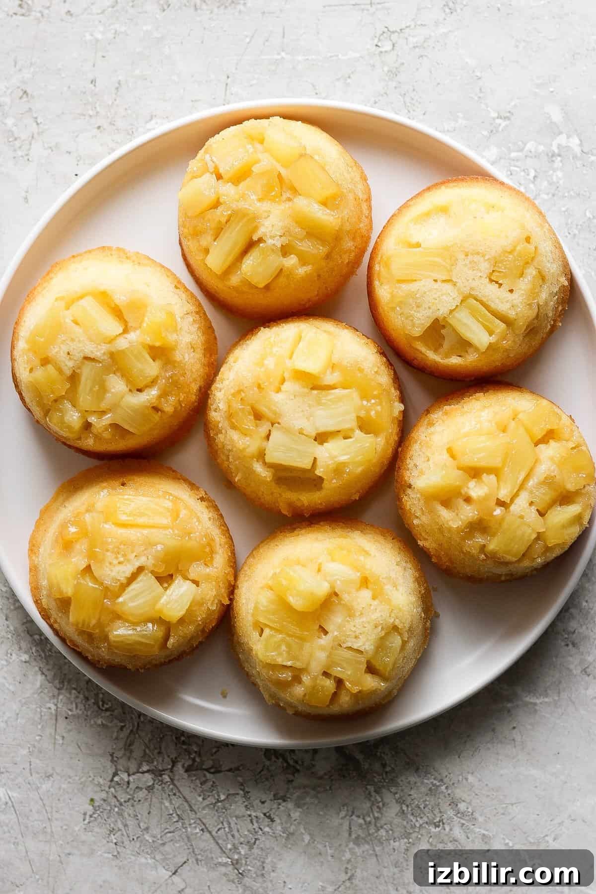 Plate of pineapple upside-down mini cakes topped with pineapple chunks on a light textured surface.