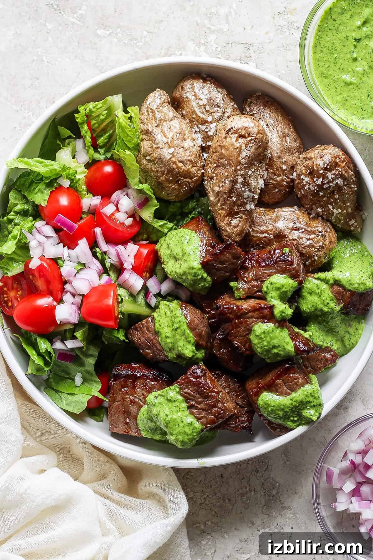 Crispy Air Fryer Steak Bites with Garlic Butter 2 A bowl of grilled steak chunks with green sauce, roasted potatoes, and a side salad of lettuce, tomatoes, and chopped red onions.