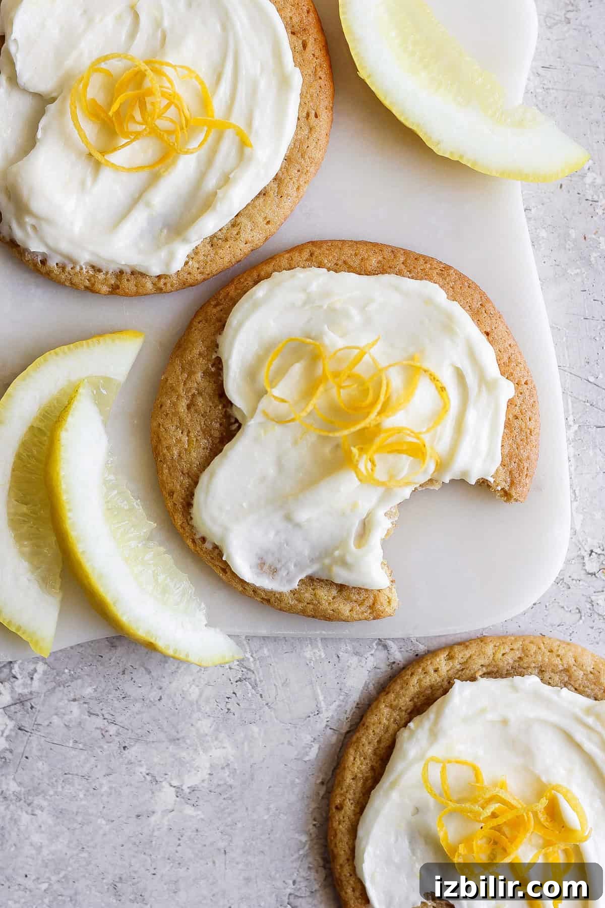 Zesty Lemon Cookies with Cream Cheese Frosting 2 Three lemon cookies with cream cheese frosting, topped with fresh zest, sit invitingly on a cutting board. One is partially eaten, revealing its soft interior. Beside them, lemon wedges are artfully arranged on the textured surface, enhancing the citrusy allure of the scene.