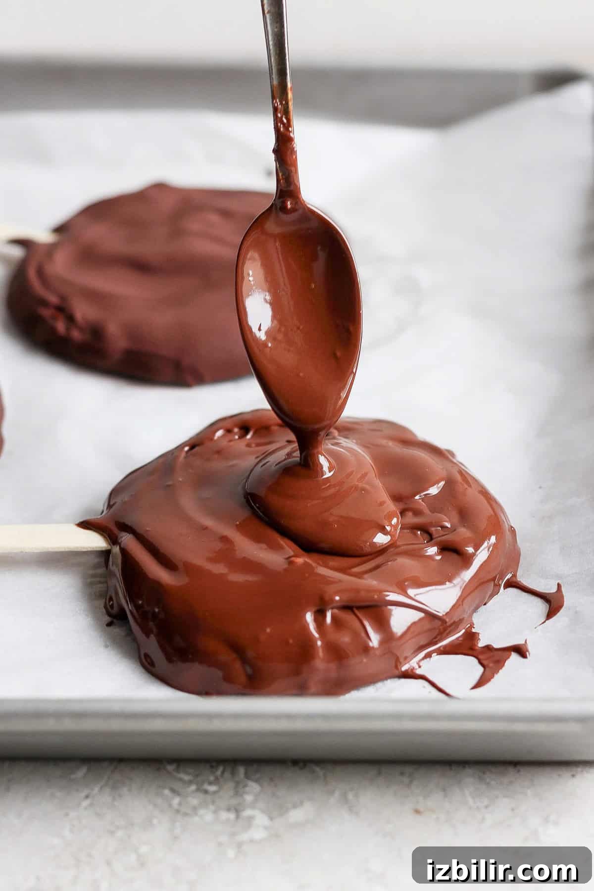 A spoon drizzles melted chocolate onto a round treat on a parchment-lined tray with a popsicle stick.