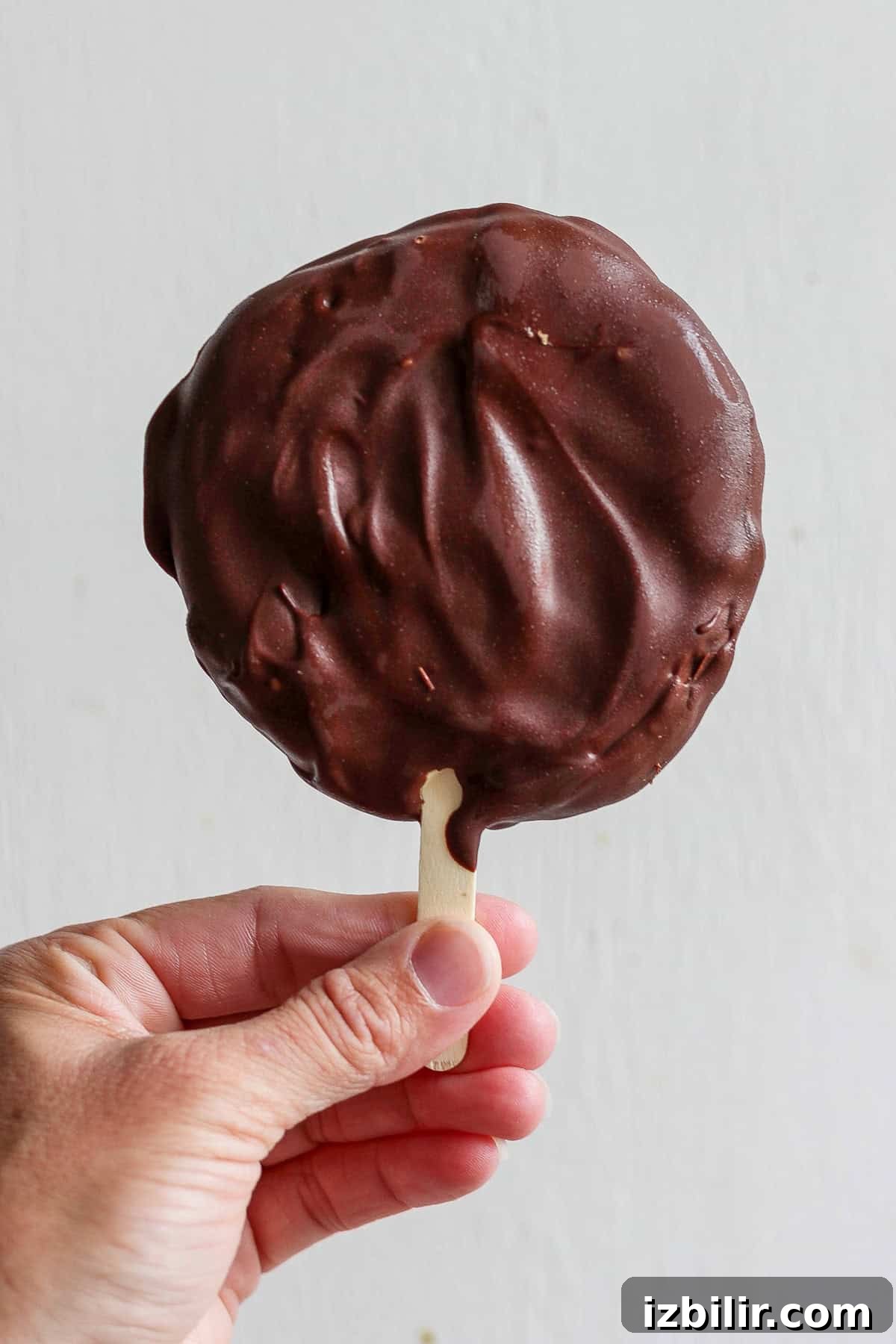 A hand holding a chocolate-covered ice cream on a stick against a plain background.