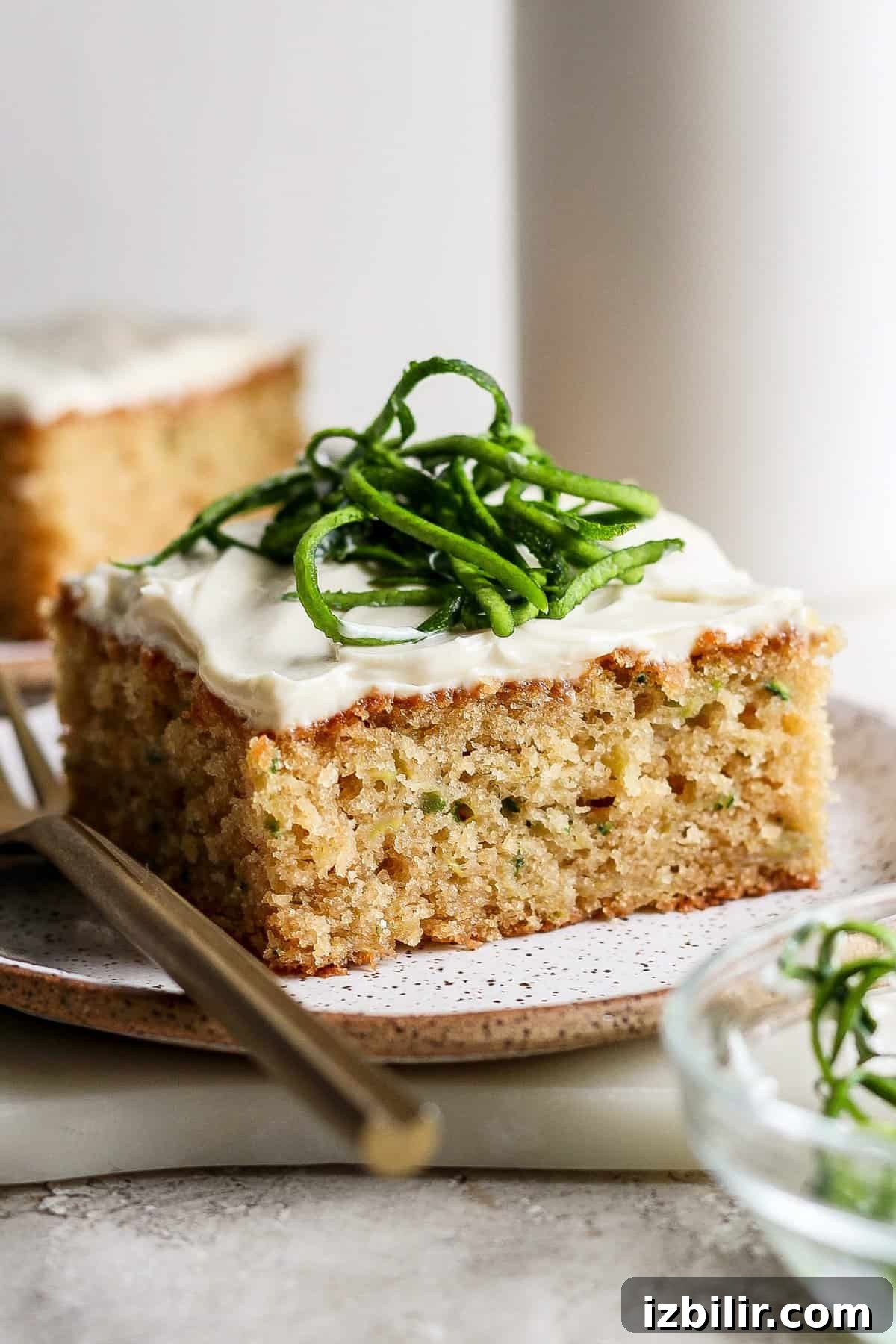 Moist Low-Sugar Zucchini Cake Recipe 2 A slice of frosted zucchini cake topped with thin slices of zucchini on a plate with a fork.
