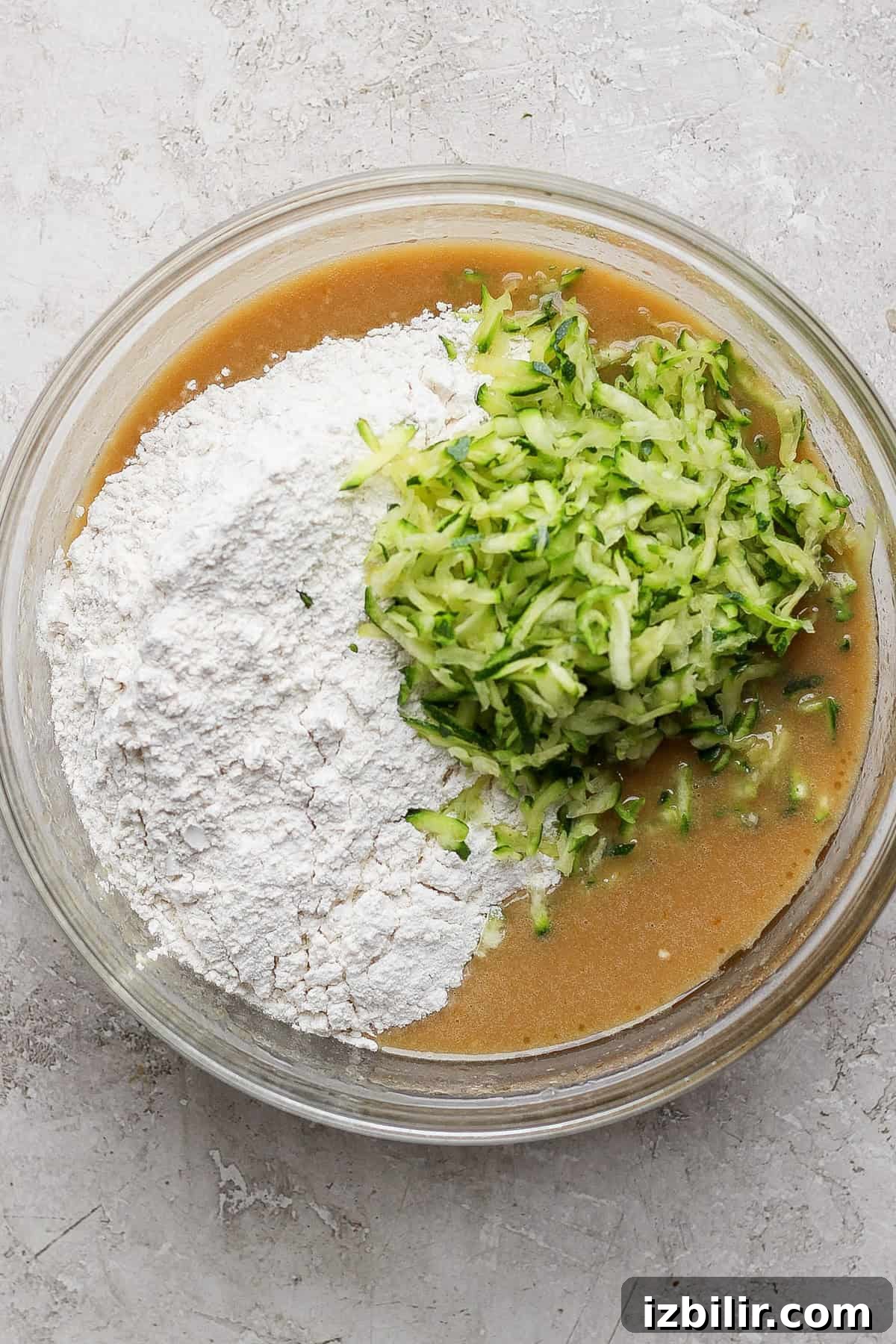 Moist Low-Sugar Zucchini Cake Recipe 3 A glass bowl containing a baking mixture with flour, brown liquid batter, and grated zucchini on a light gray surface.